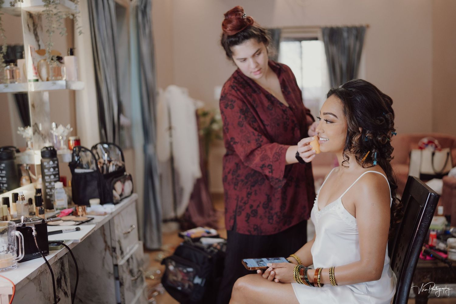 A stylist works on a bride's makeup at a wedding prep vanity station with beauty products and tools nearby.