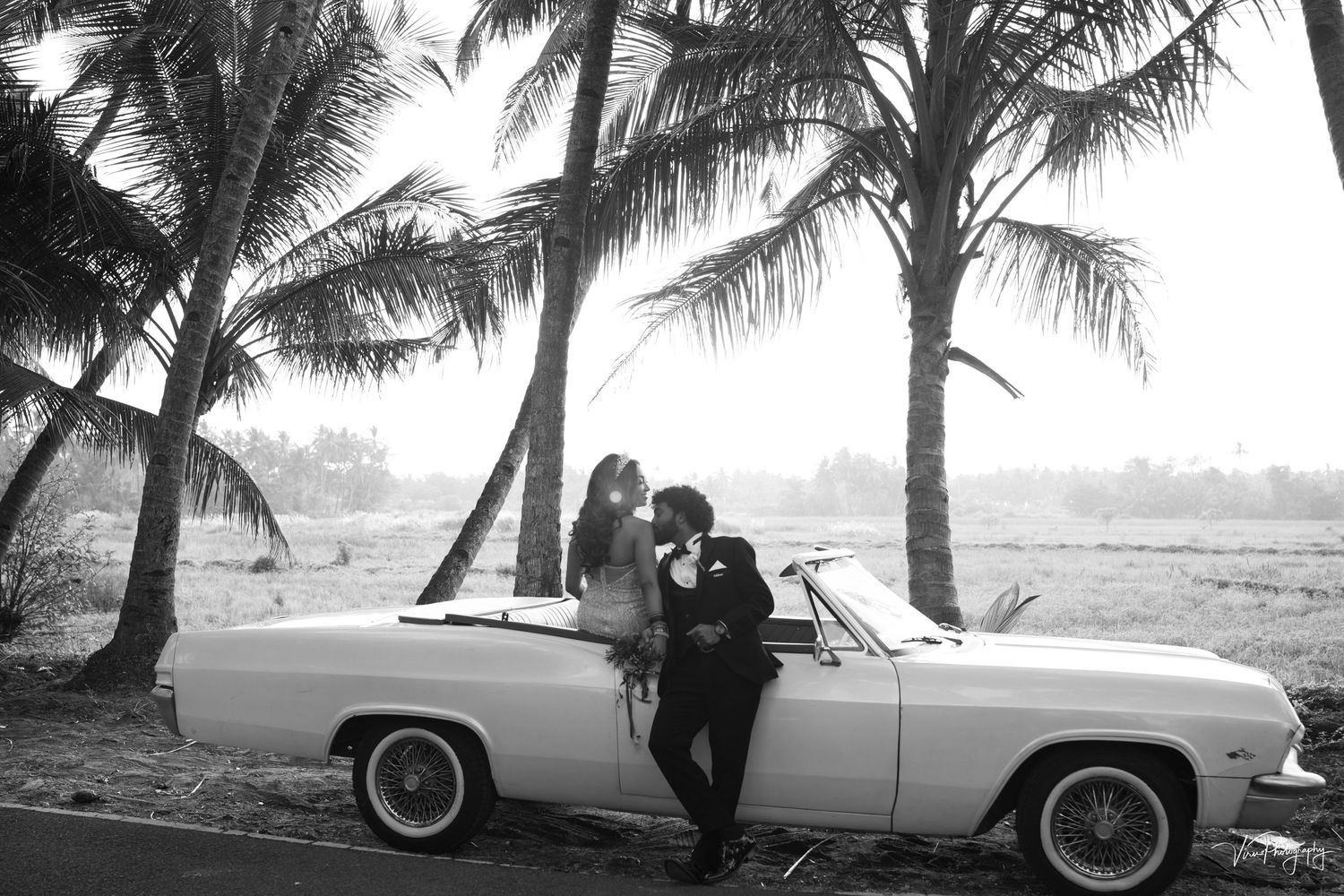 Classic convertible car parked beneath palm trees along a tropical beach in vintage black and white photograph.