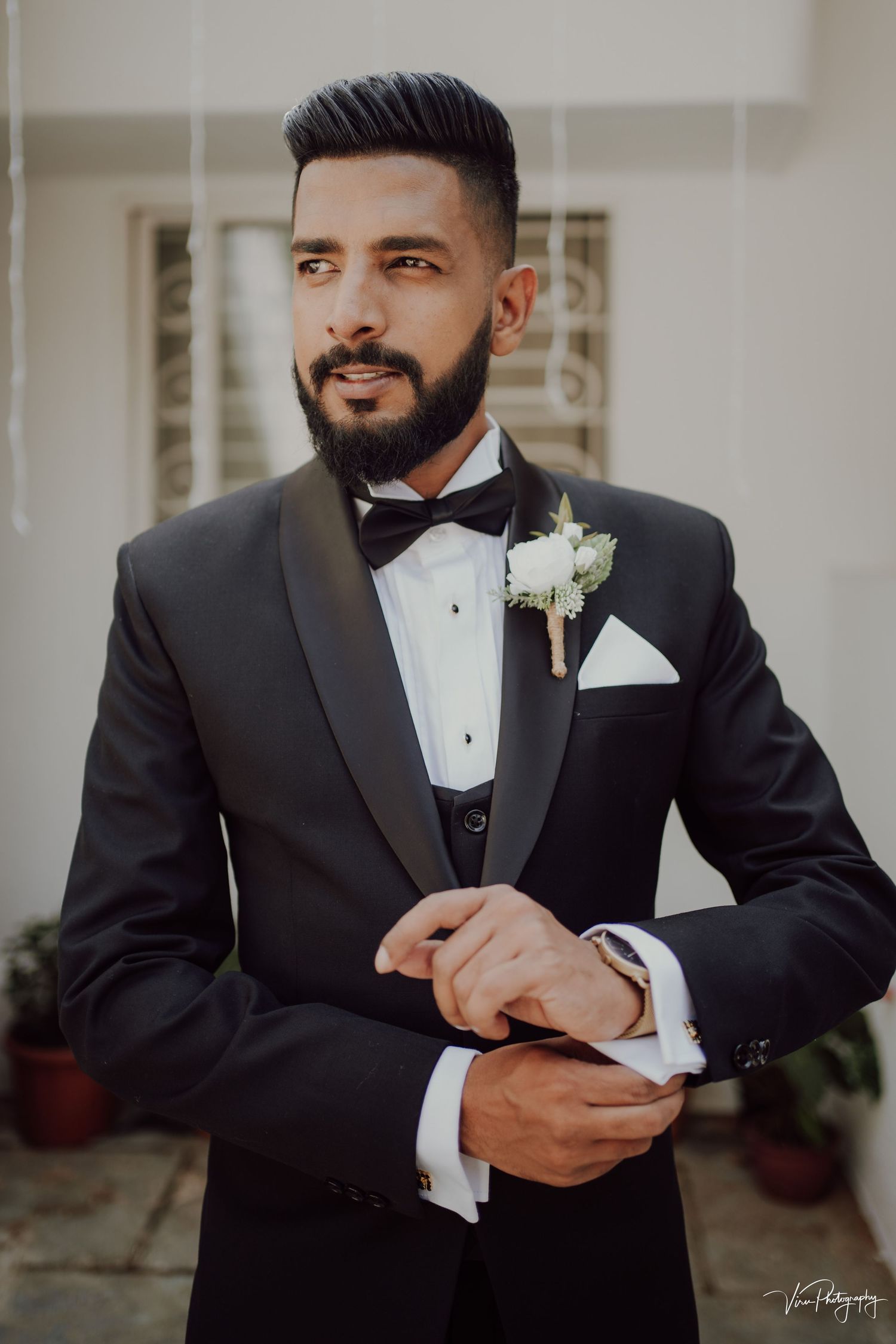 Groom in classic black tuxedo with bow tie and white boutonniere adjusts cufflinks on wedding day.