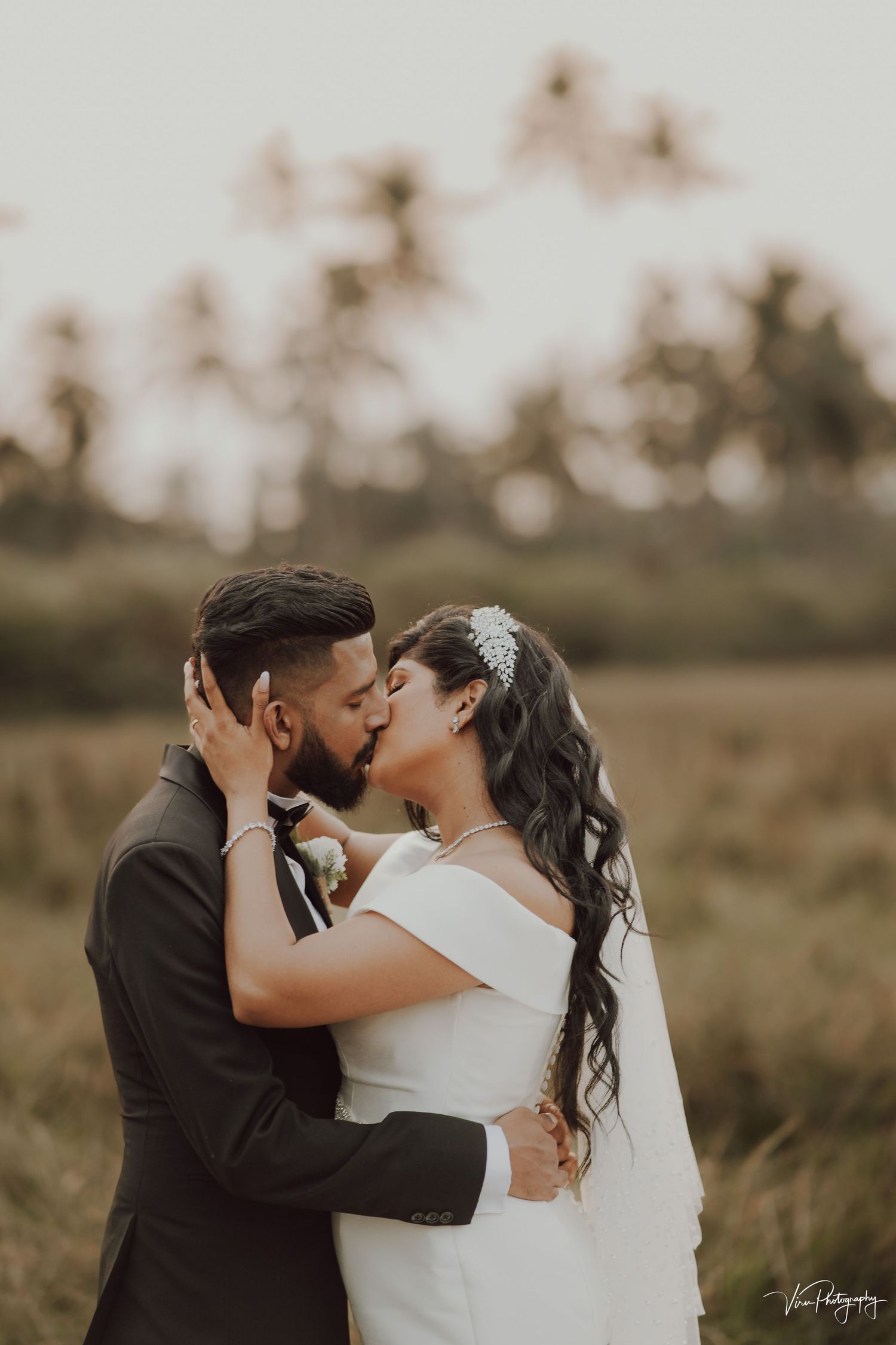 A romantic outdoor wedding photo of a couple sharing a tender kiss in a rustic field at sunset.