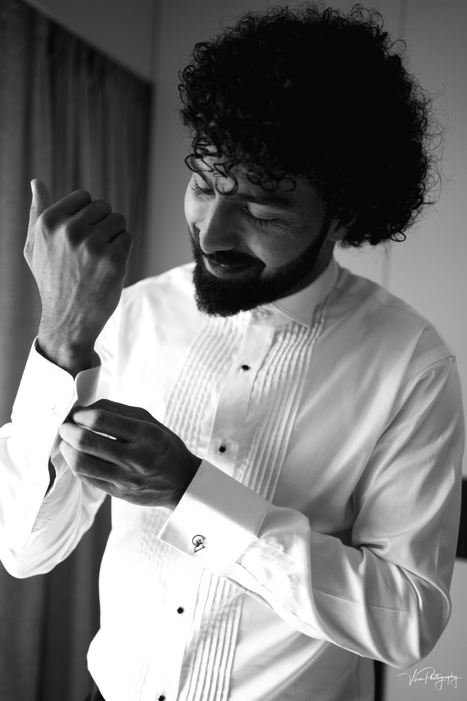 Man in white dress shirt with curly dark hair and beard adjusts his cuff links while getting ready.
