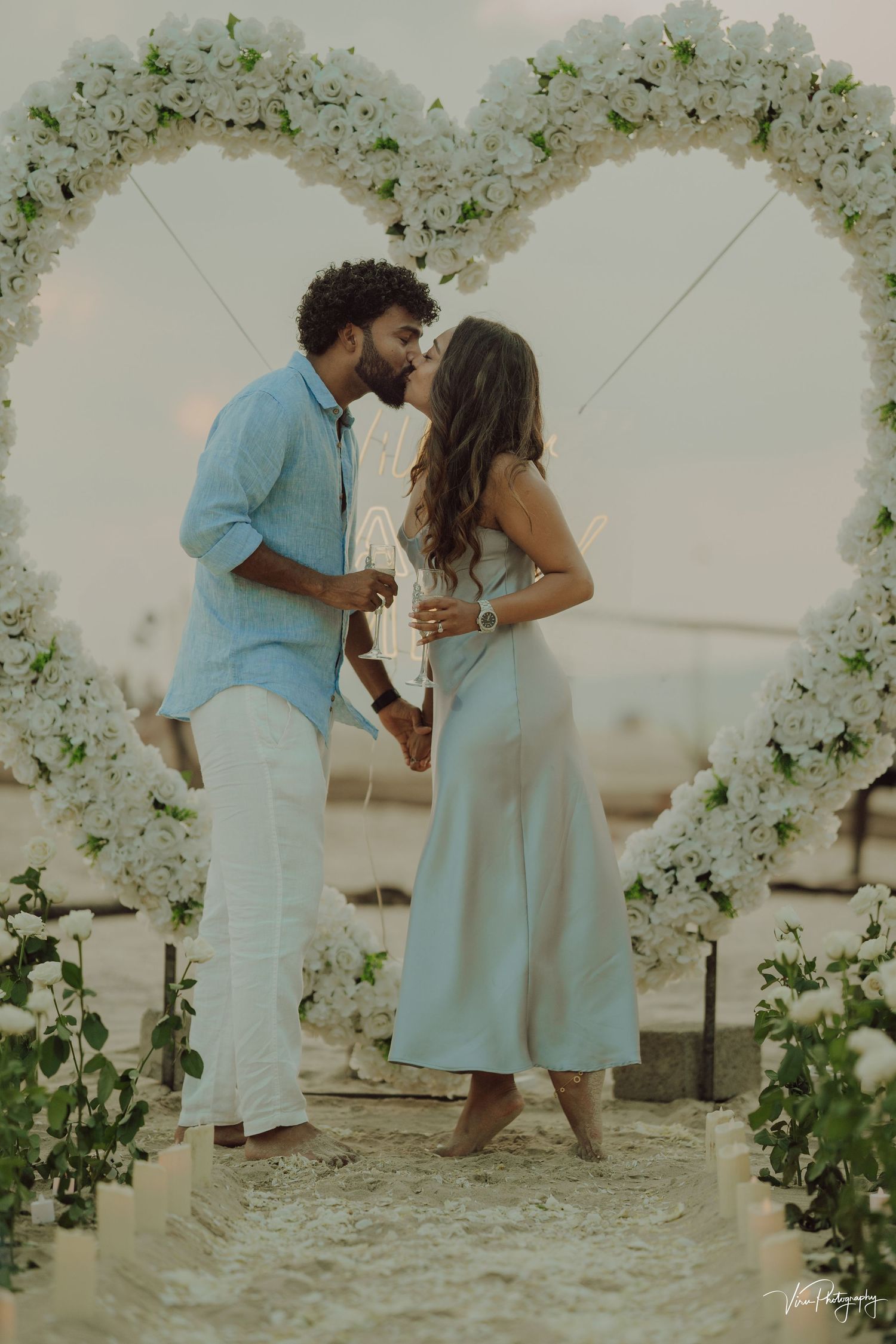 A couple shares a romantic moment under a white floral arch at a beach wedding ceremony at sunset.