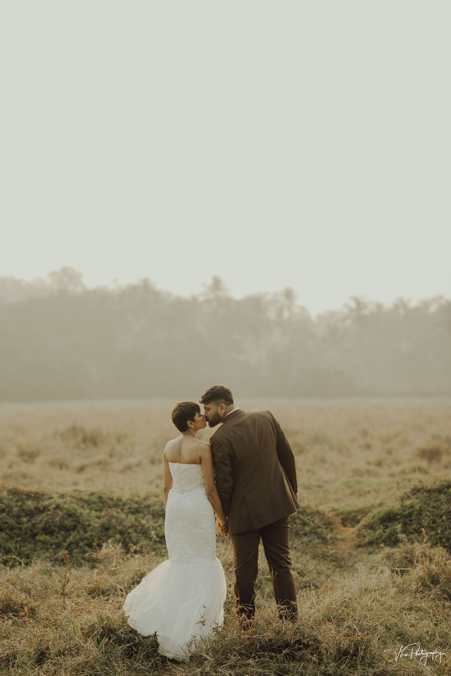 A couple in wedding attire walks together through a misty outdoor field at dusk creating a romantic silhouette.