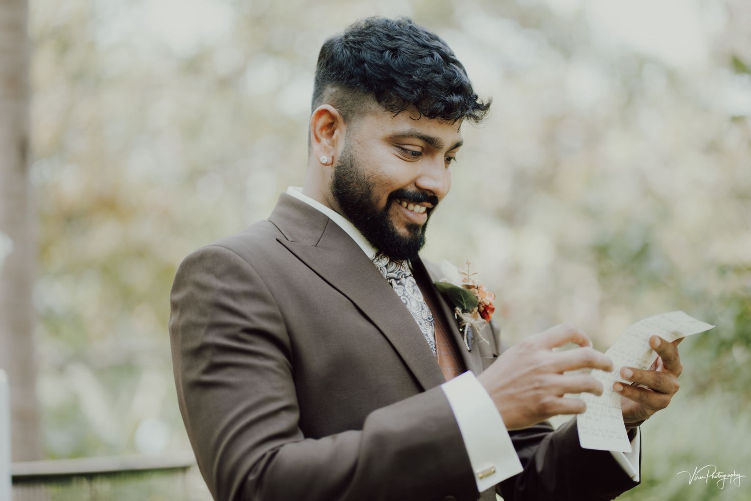 Groom in brown suit smiles while looking at cell phone during outdoor wedding ceremony.