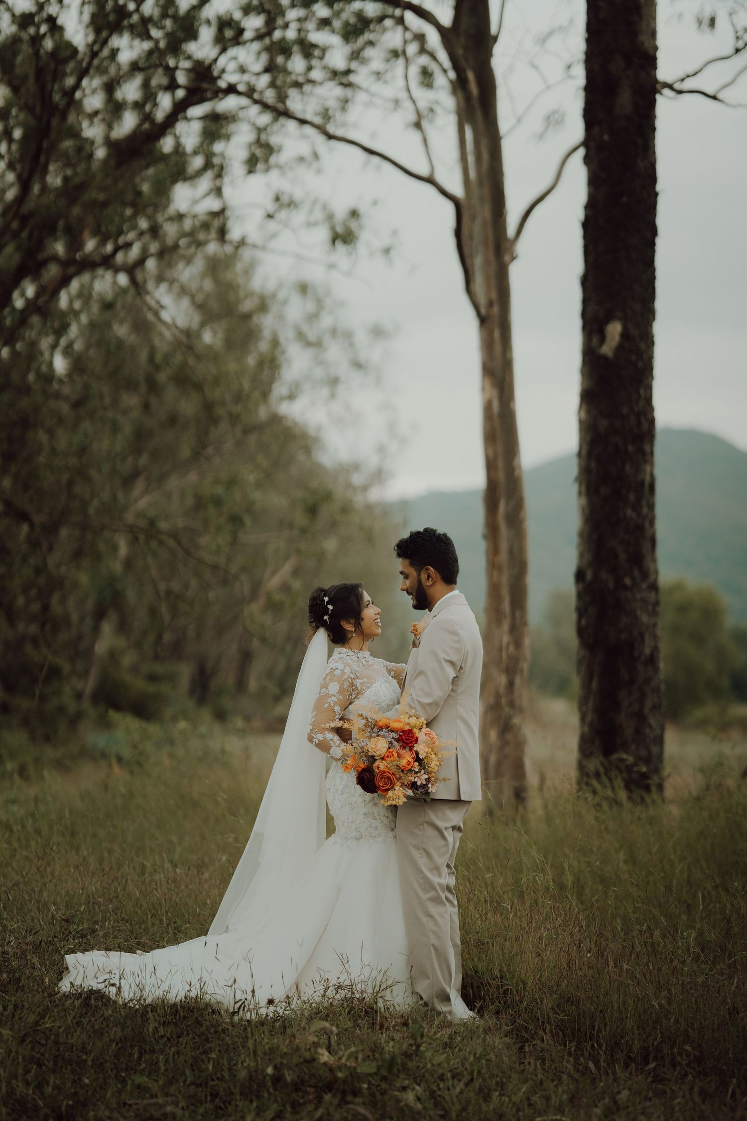 A bride in a long veil and groom in a beige suit embrace in a grassy field with tall trees and misty mountains behind them.