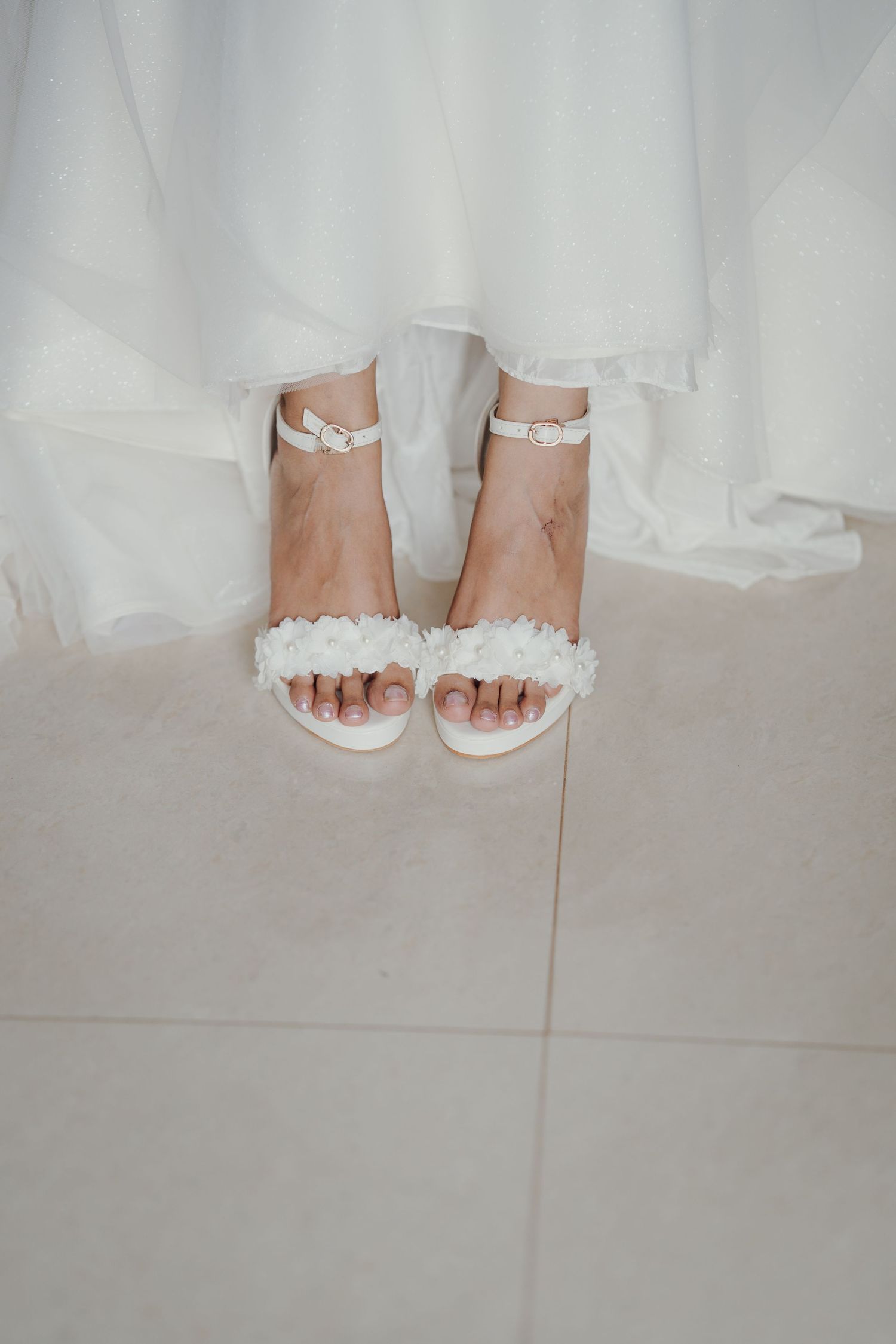 Bride's feet in white block-heel sandals with floral appliqué straps standing on a tiled floor beneath a flowing white gown.