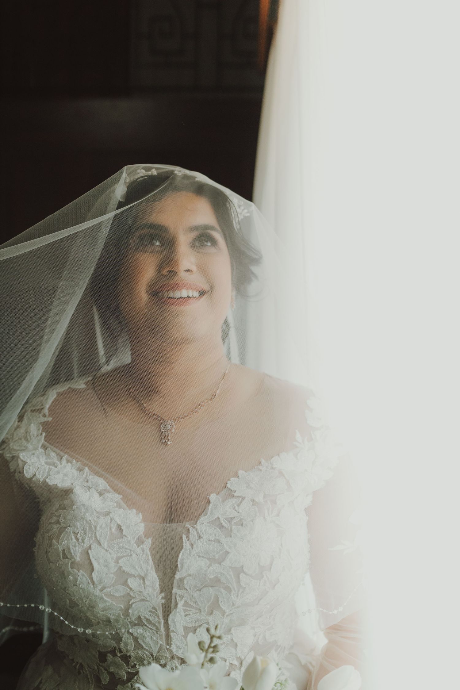 Bride wearing an elegant lace wedding dress and veil smiles radiantly in natural light.