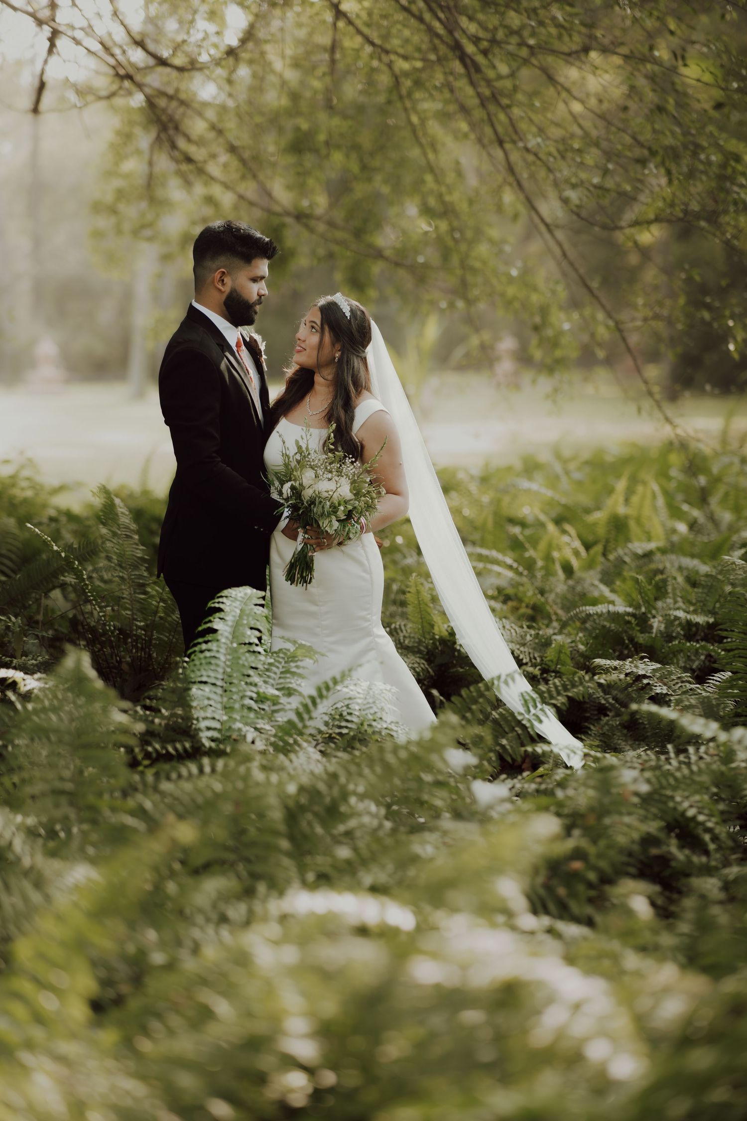 A couple embraces in their wedding attire surrounded by lush ferns and greenery on a misty forest path.