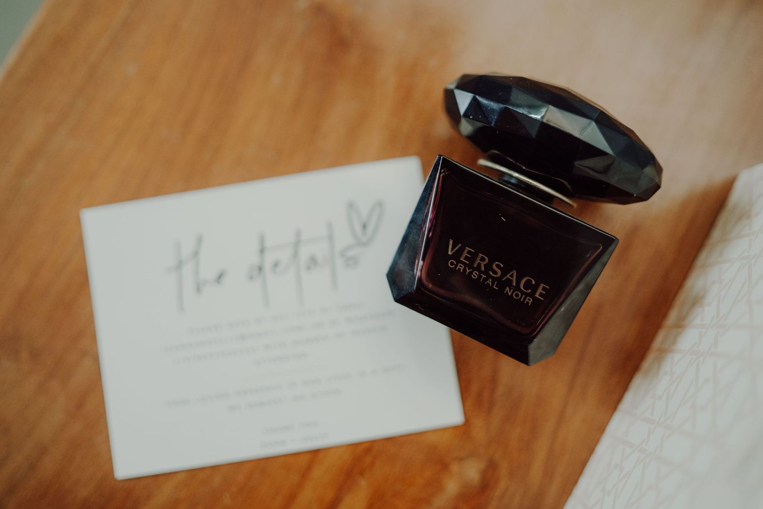 A black Versace perfume bottle sits next to a wedding invitation on a wooden surface.