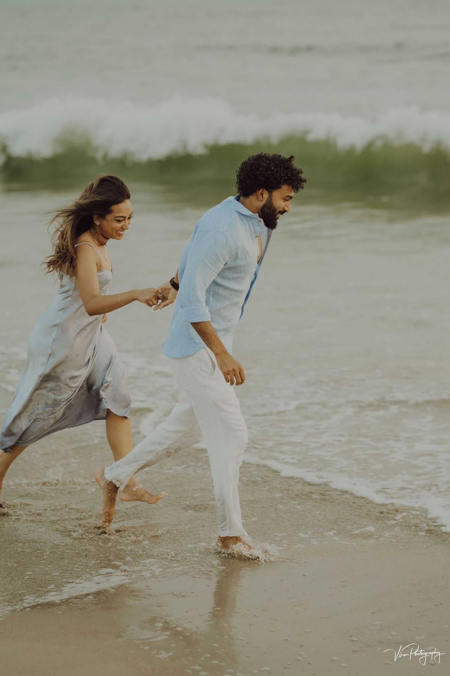 Couple joyfully running along the beach shoreline wearing light colored casual clothes at dusk.
