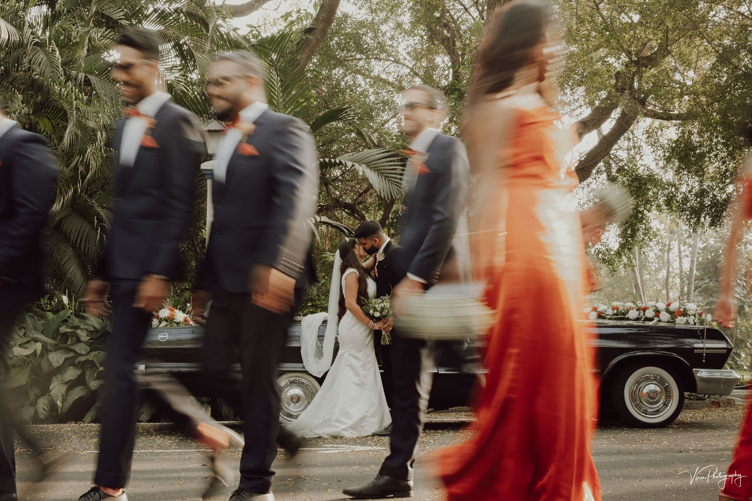 Motion-blurred figures in formal attire walking past vintage cars at an outdoor event with natural greenery.