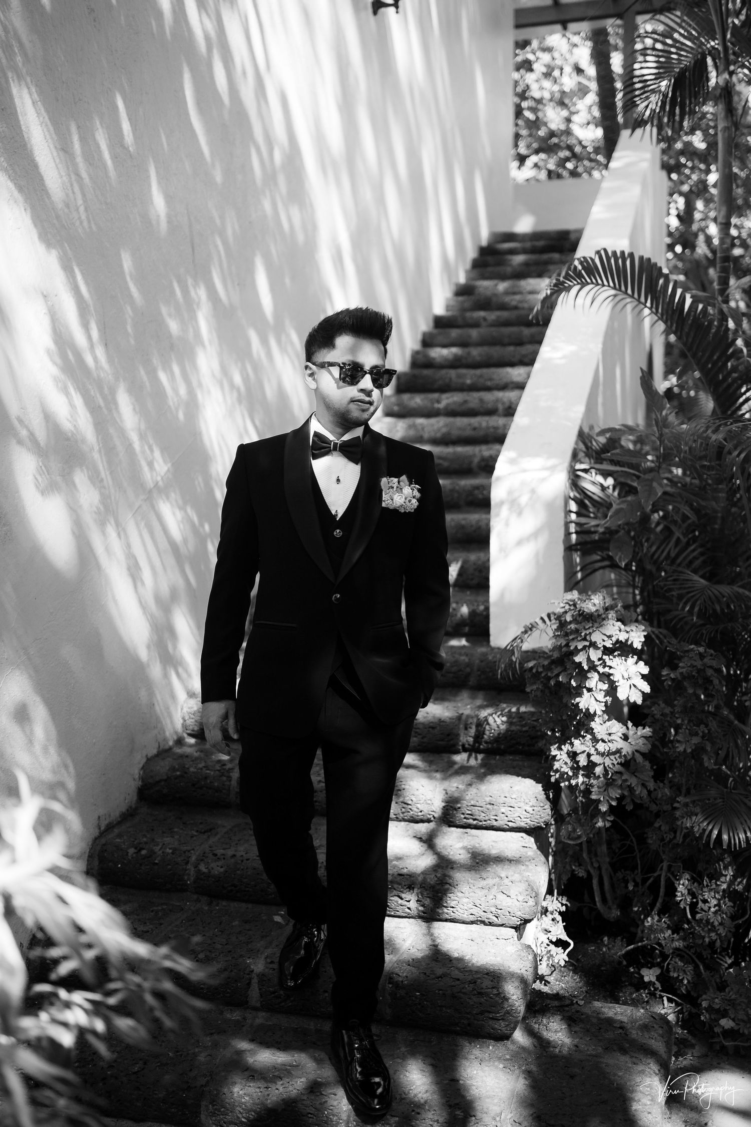 A black and white photo shows someone in a formal tuxedo with sunglasses standing on outdoor stone steps.