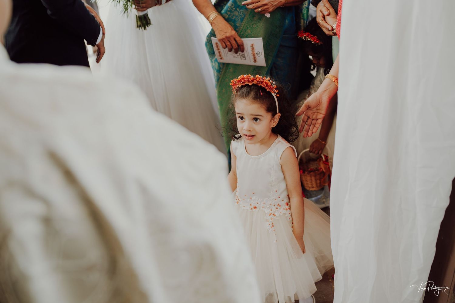 A young flower girl in a white dress stands among wedding guests during the celebration.
