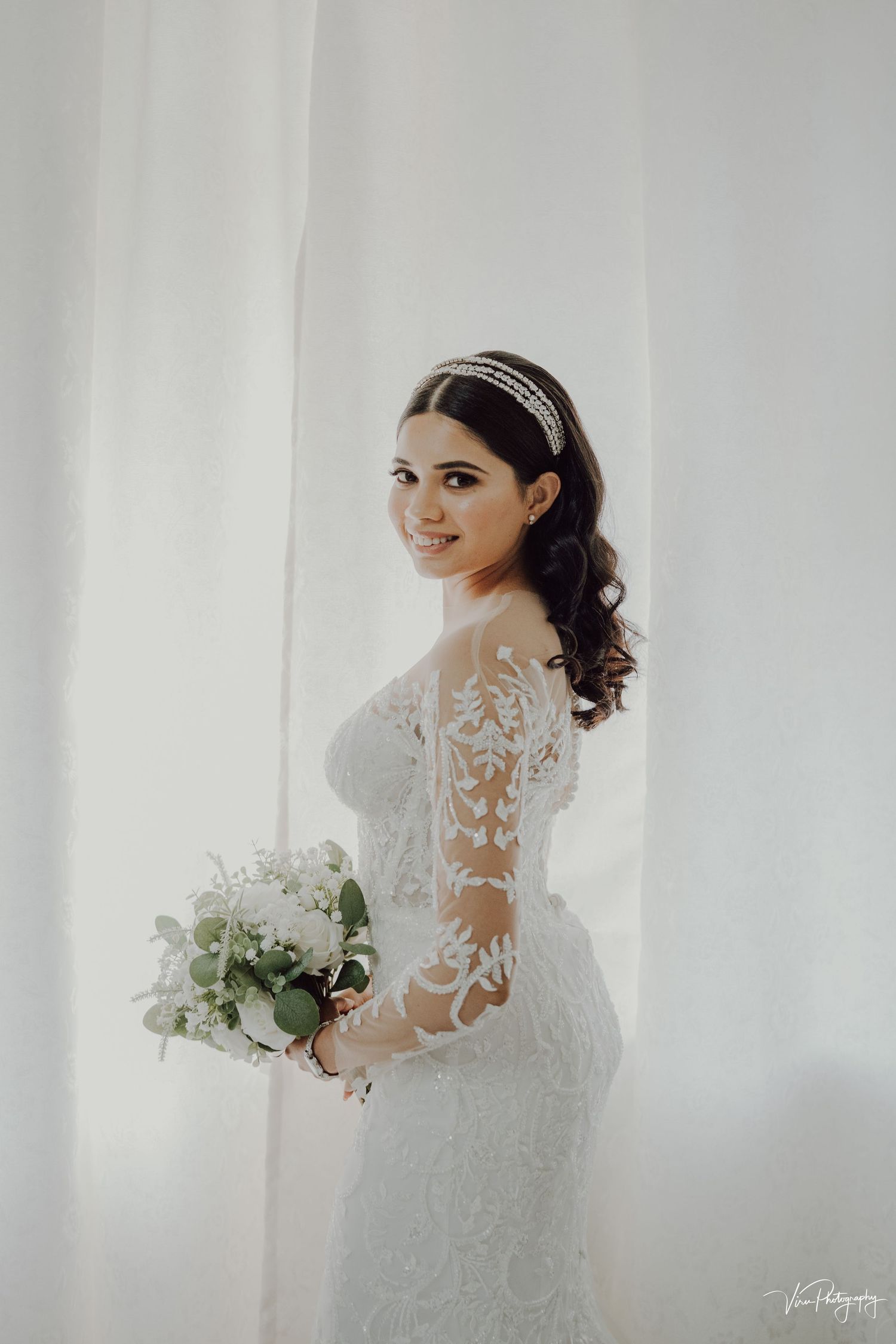 Bride in elegant lace wedding gown with sheer sleeves holds white and green bouquet against light curtained backdrop.