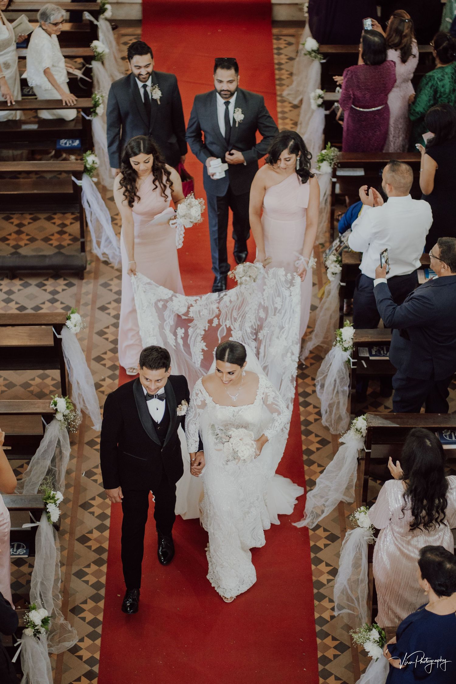 Couple walks down red carpet aisle in church during wedding ceremony with pampas grass floral decorations lining the path.