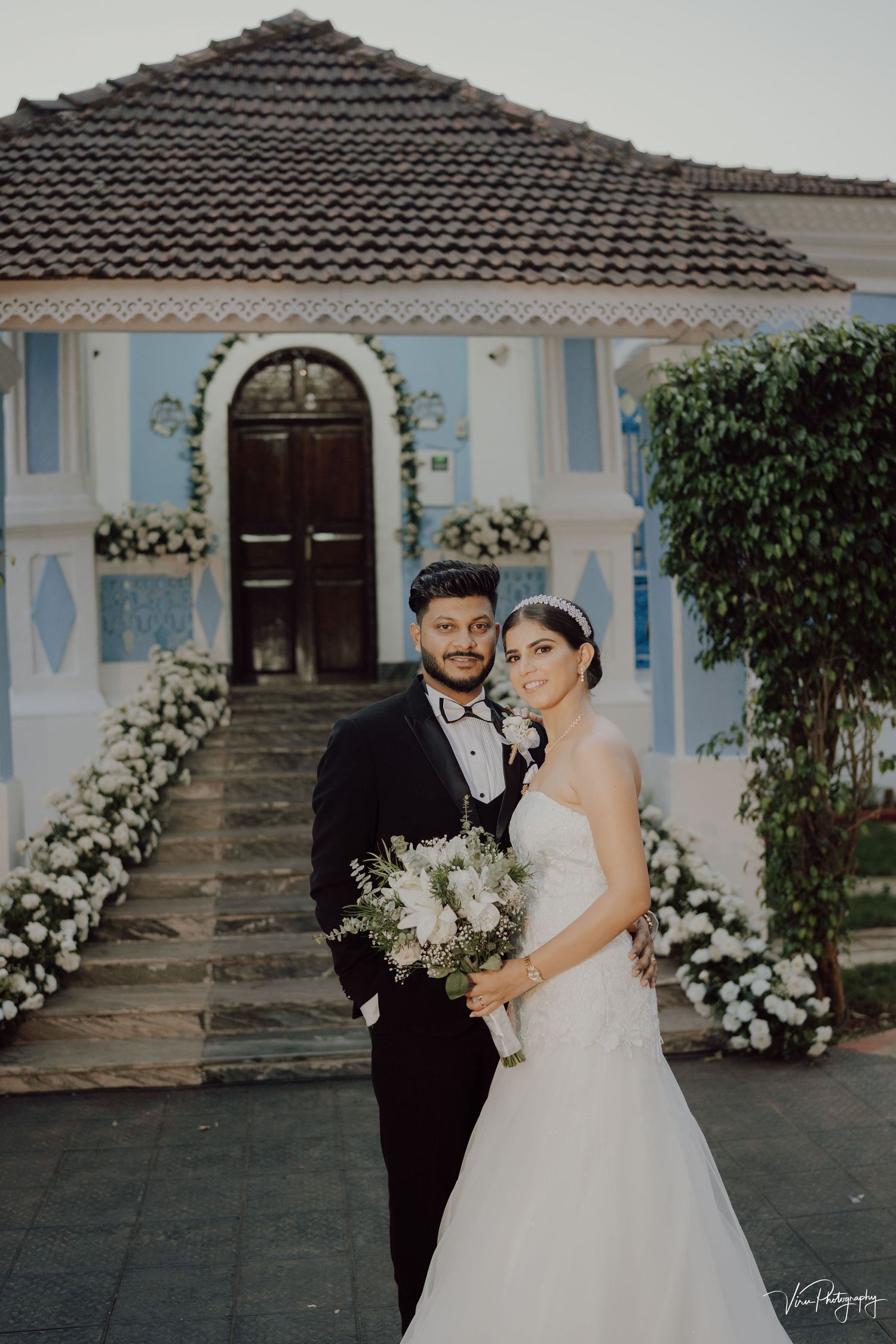 A wedding couple stands in front of a charming blue and white historic house with floral arch and tiled roof.