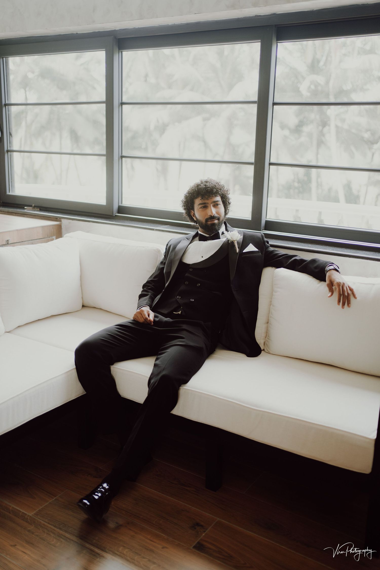A person in formal attire lounges on a white sectional sofa near large windows with natural light streaming through.