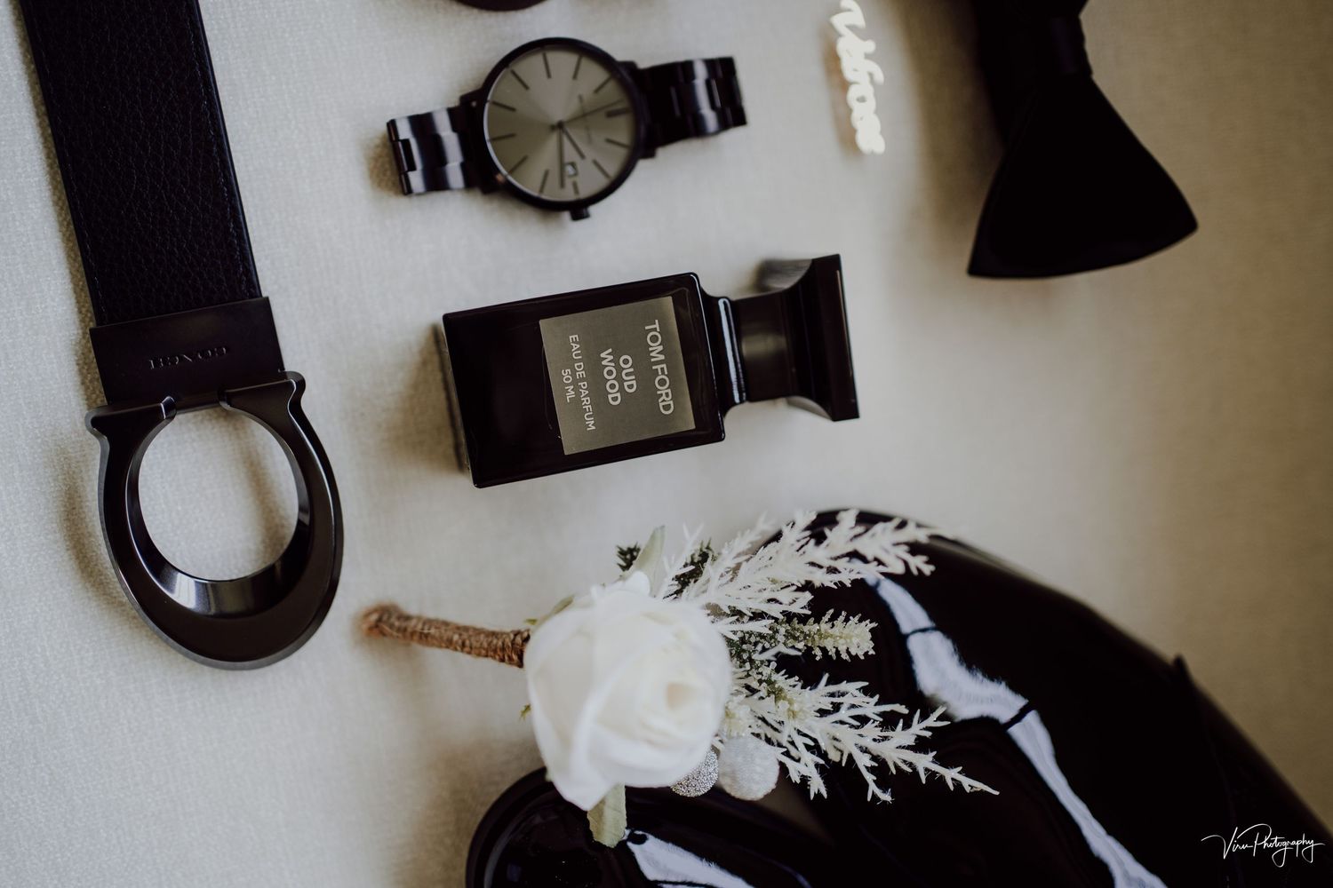 Wedding accessories including a black wrist watch, perfume bottle, and white boutonniere arranged on white surface.