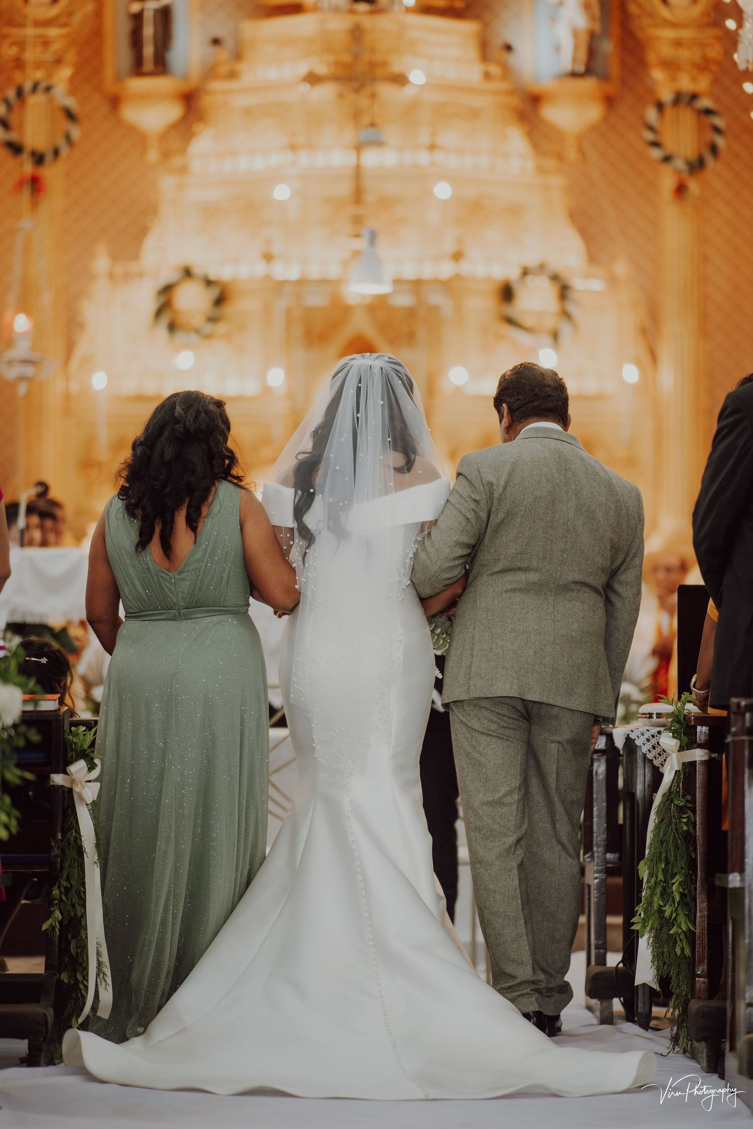 A wedding party stands at the altar in a golden-lit church with the bride wearing a white mermaid gown and long veil.