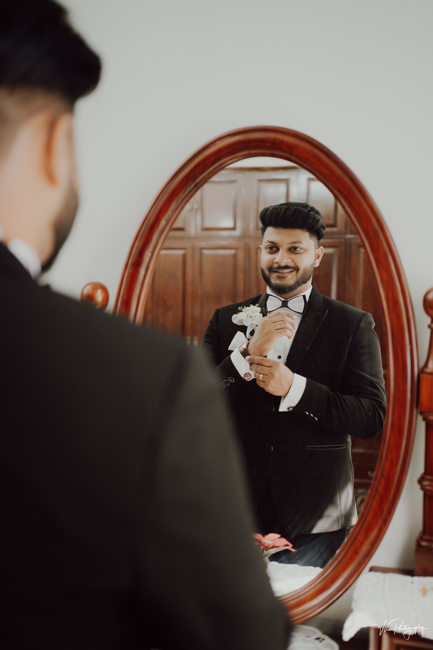 Reflection in oval vintage mirror shows groom in black tuxedo and bow tie adjusting his outfit before wedding ceremony.
