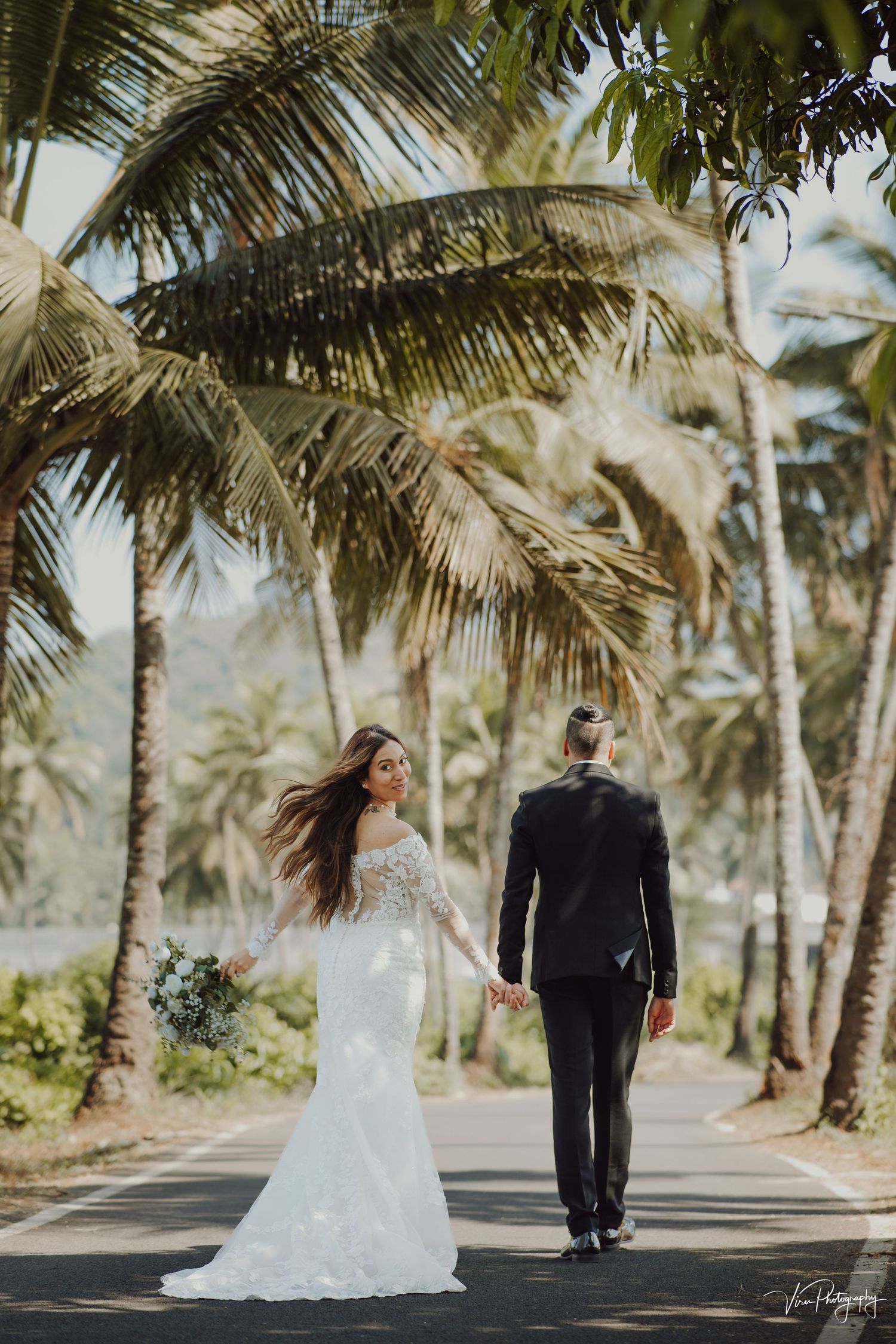 A couple in wedding attire walking down a palm tree-lined road in a tropical destination.
