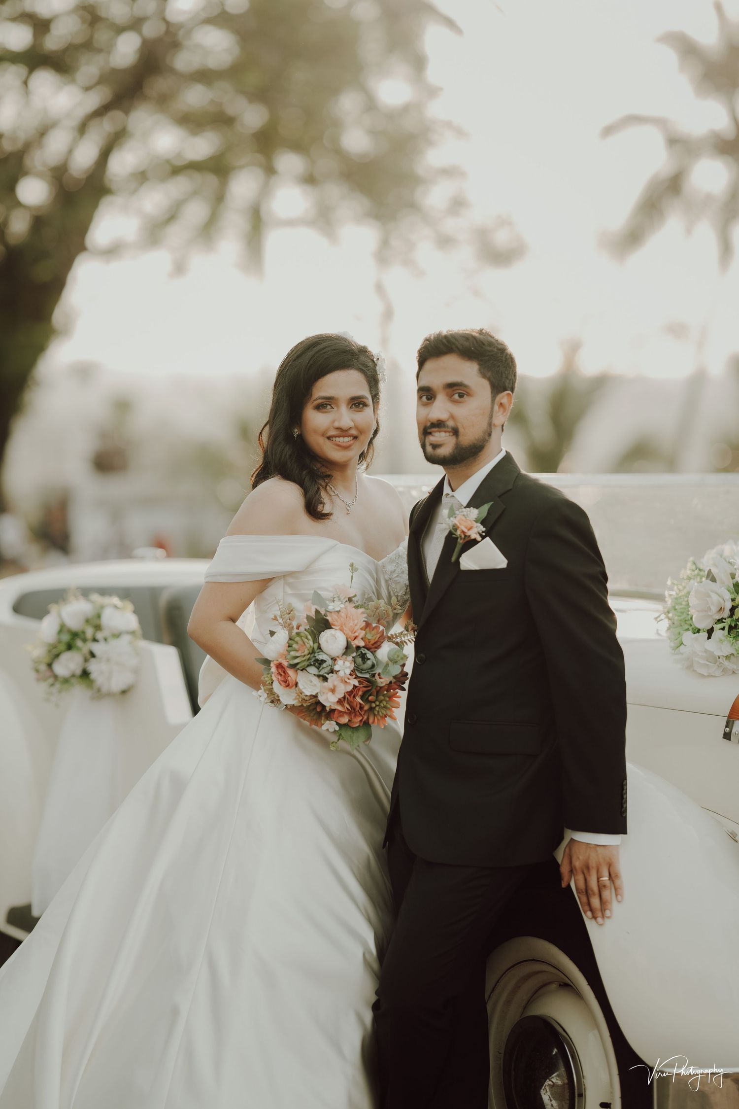 A bride in a white off-shoulder gown and groom in black suit pose together outdoors with a romantic floral bouquet.