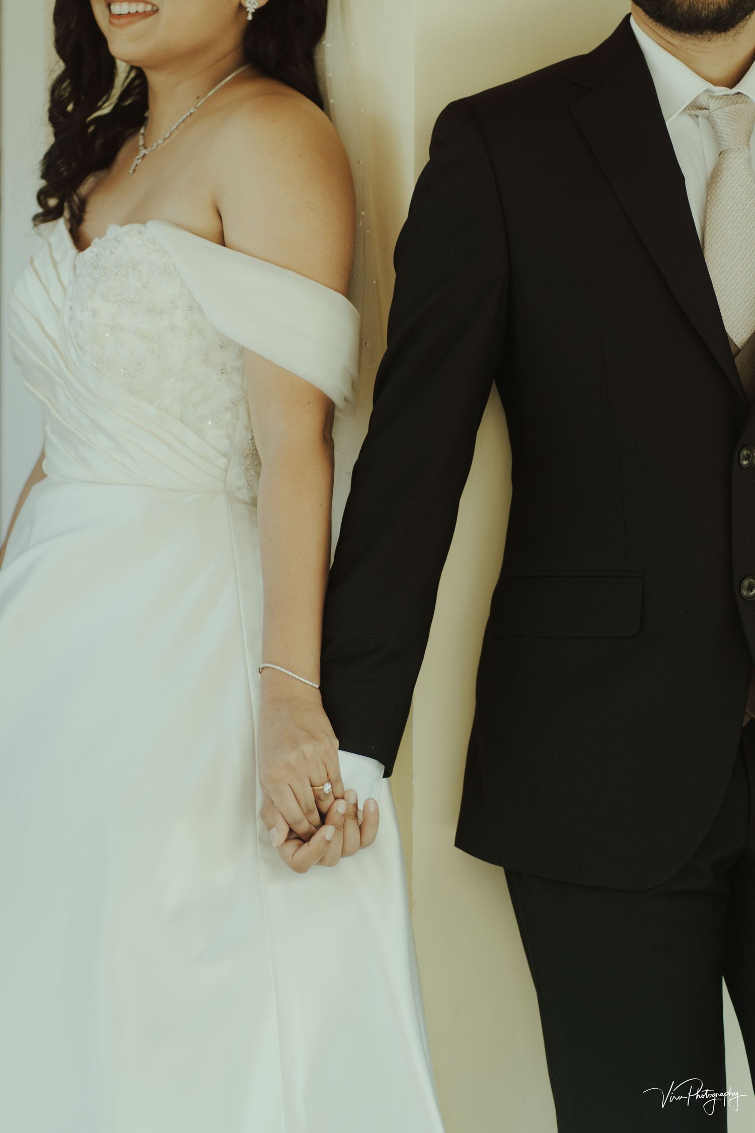 A close-up of a bride and groom holding hands, showing her off-shoulder white gown and his classic black suit.