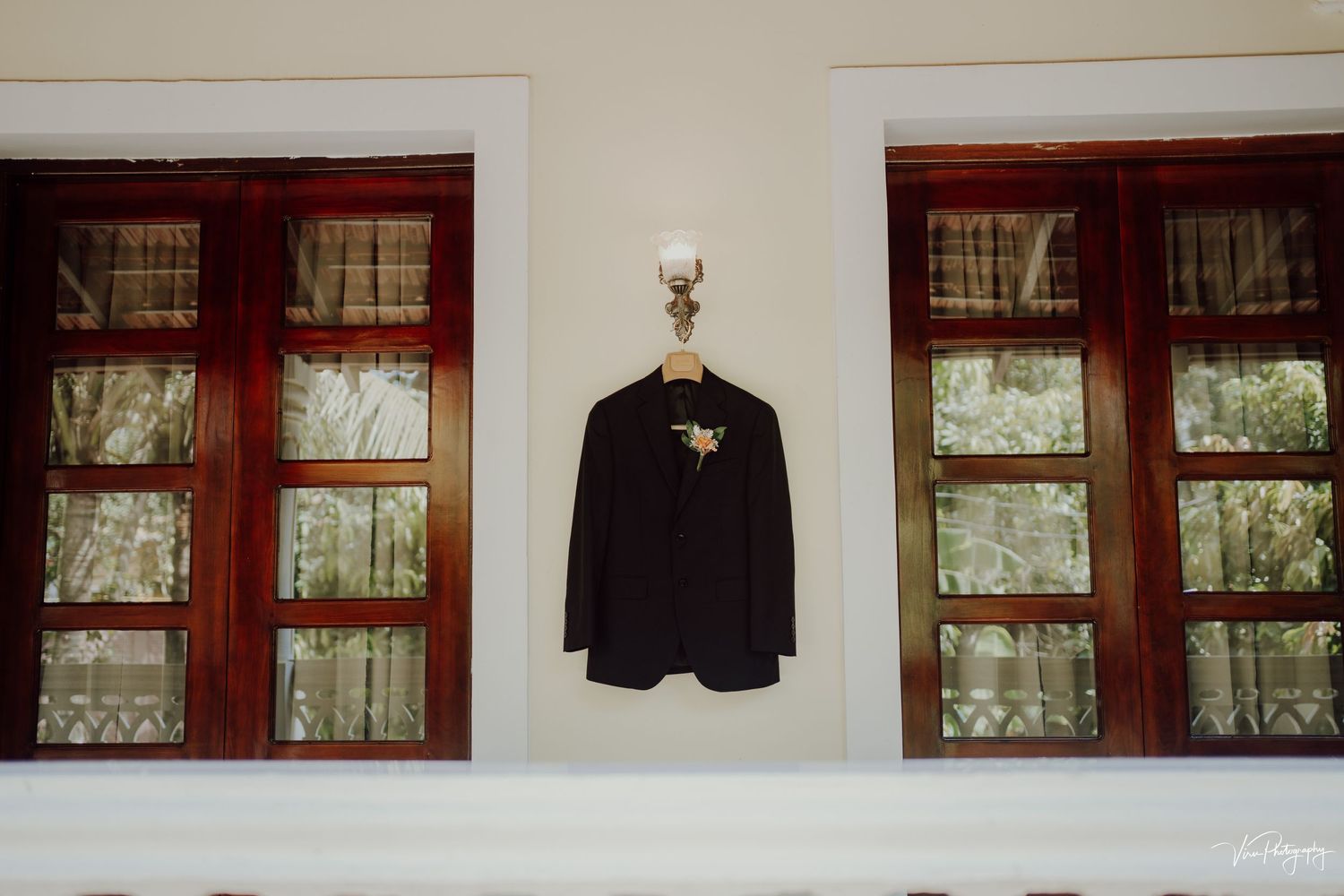A black suit jacket hangs on a wall between two red-framed windows with sheer curtains.