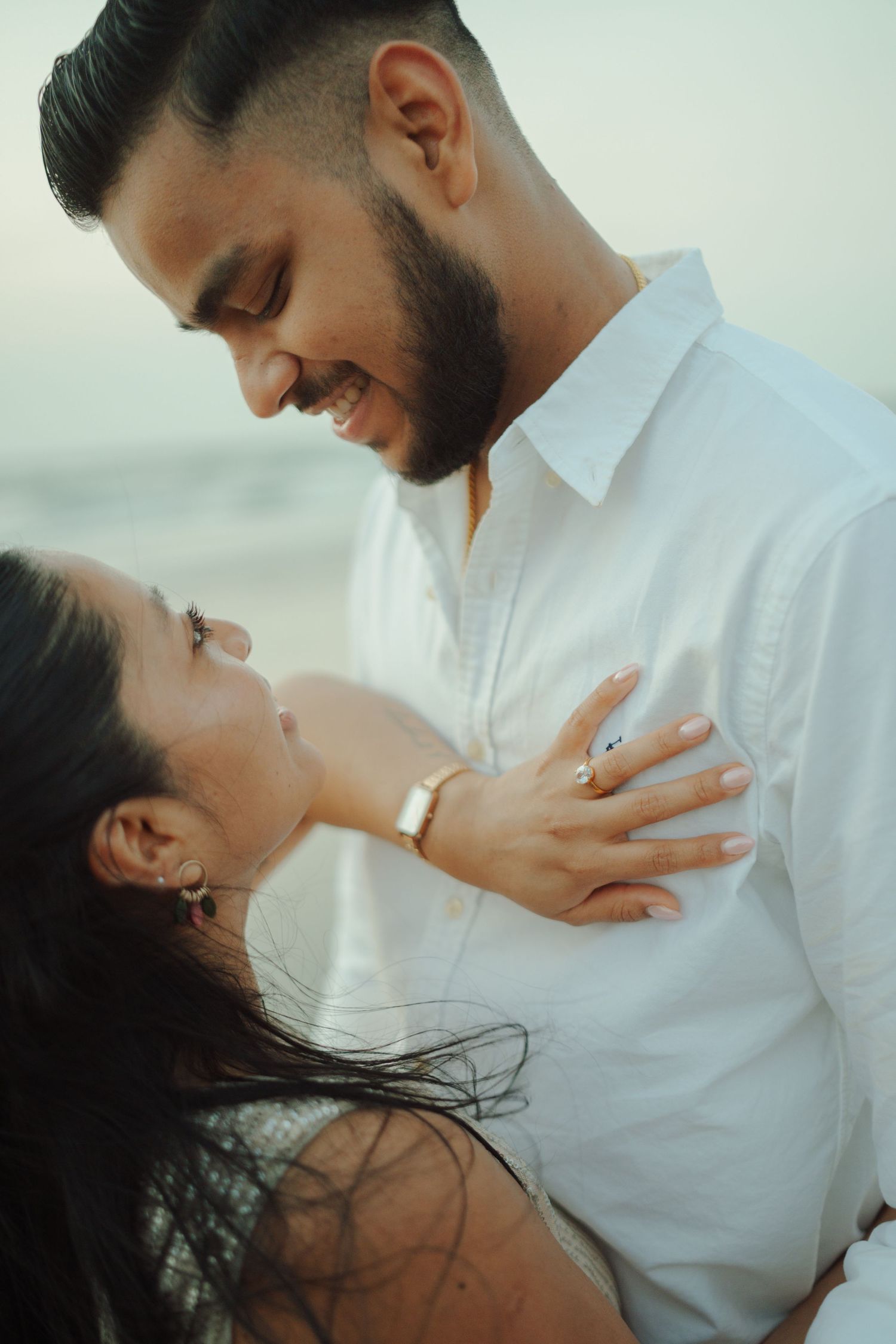 A smiling couple embracing on a beach, the man in a white shirt gazing down at the woman who looks up at him lovingly.