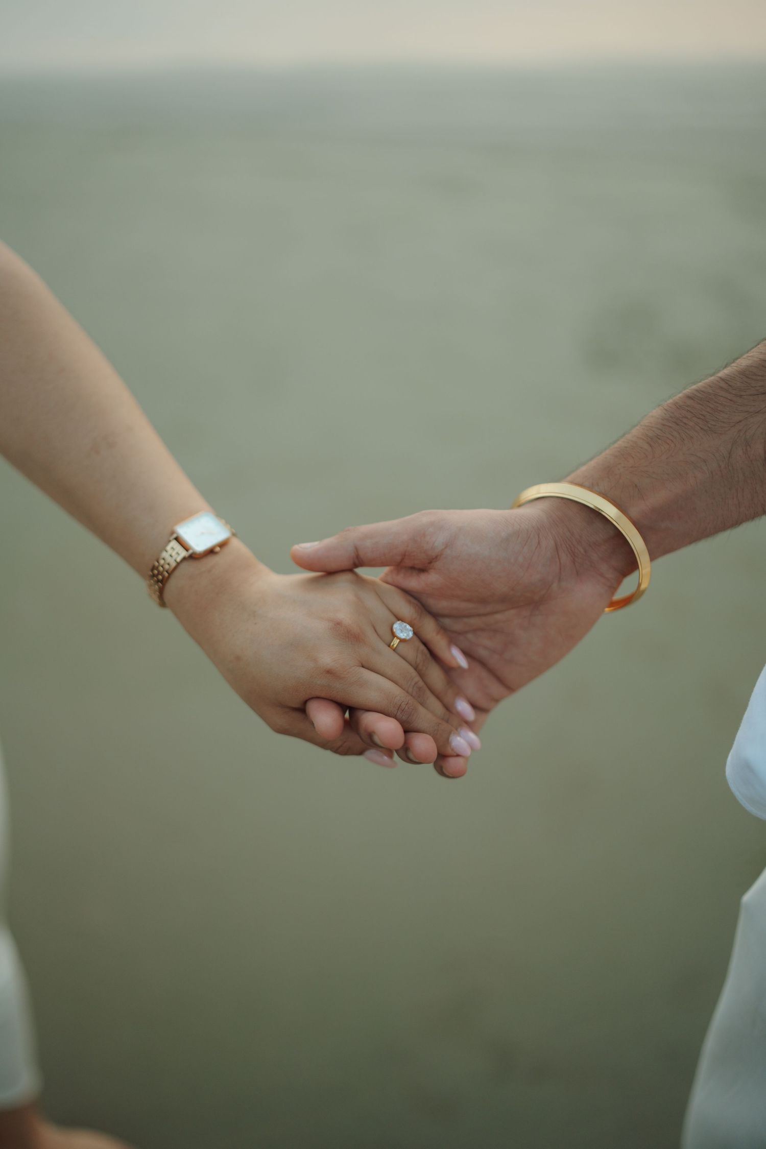 Couple holding hands on beach, woman wearing diamond ring, gold watch, and bracelet, man wearing gold bangle.
