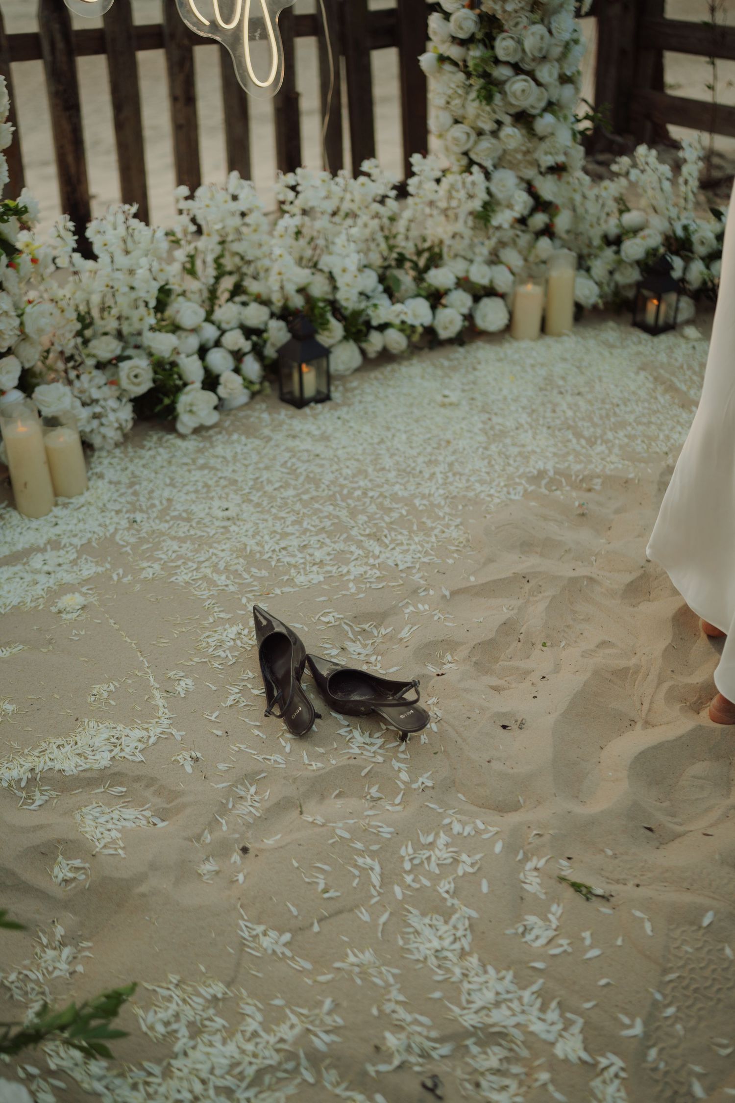 Dark heels resting on sandy beach wedding aisle with white petals, candles, and floral arrangements in background.