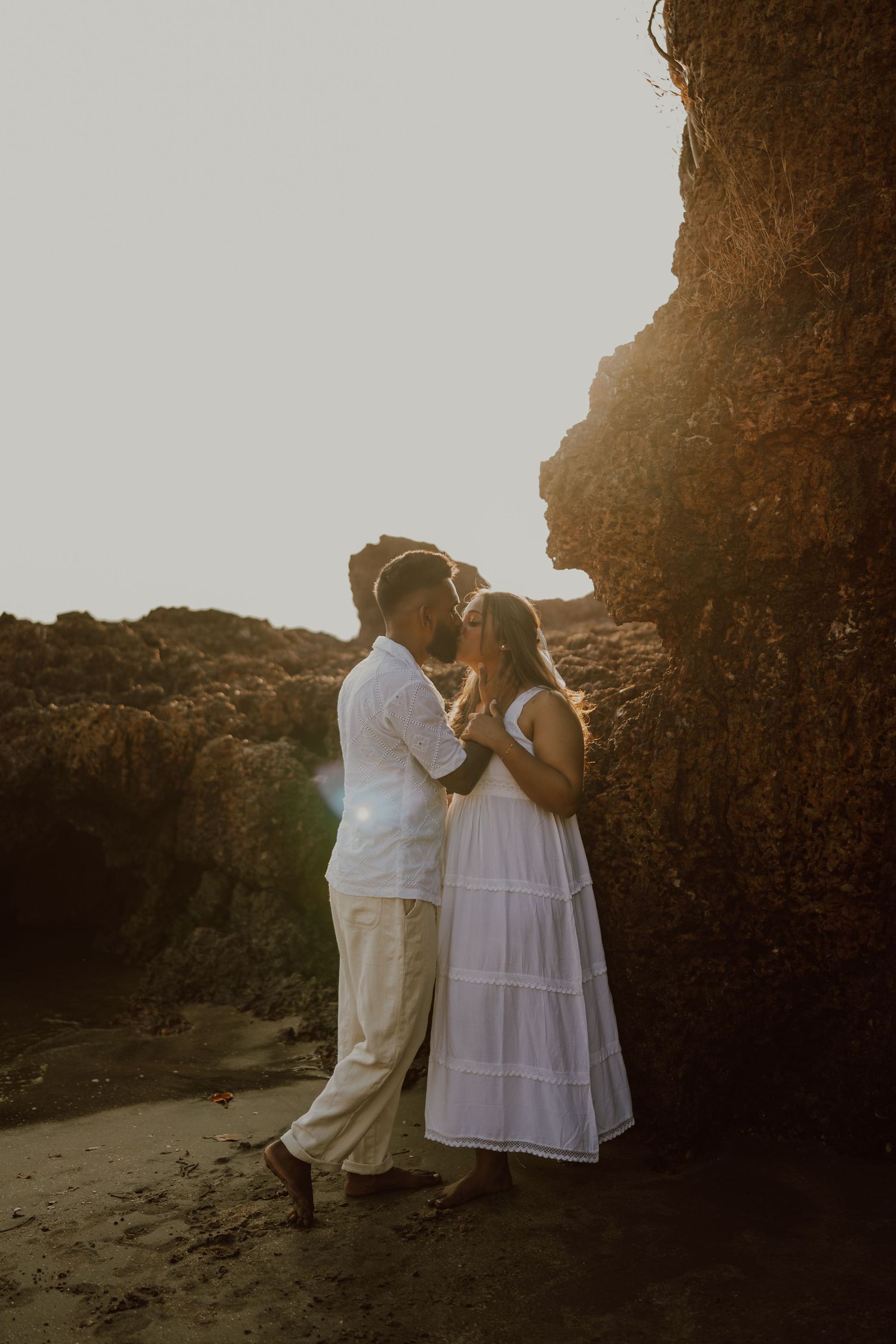 A couple in white outfits shares an intimate moment against a rocky beach backdrop at sunset.