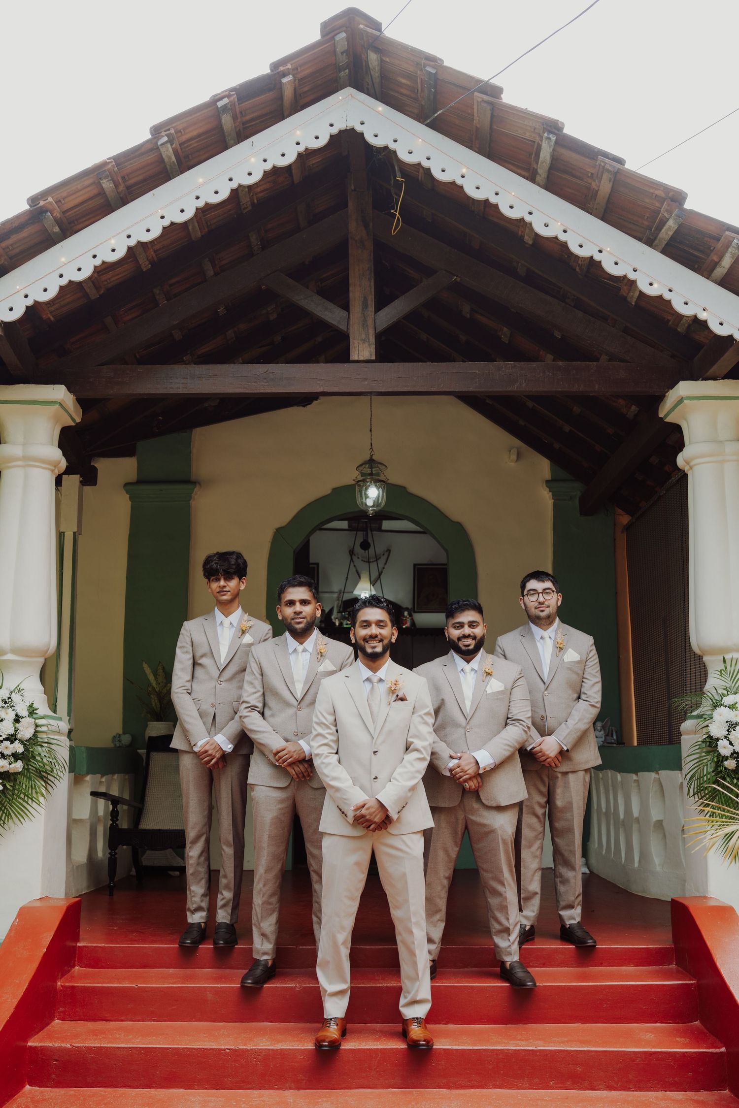 Five groomsmen in beige suits stand on red steps in front of a traditional tiled-roof building decorated with white flowers.