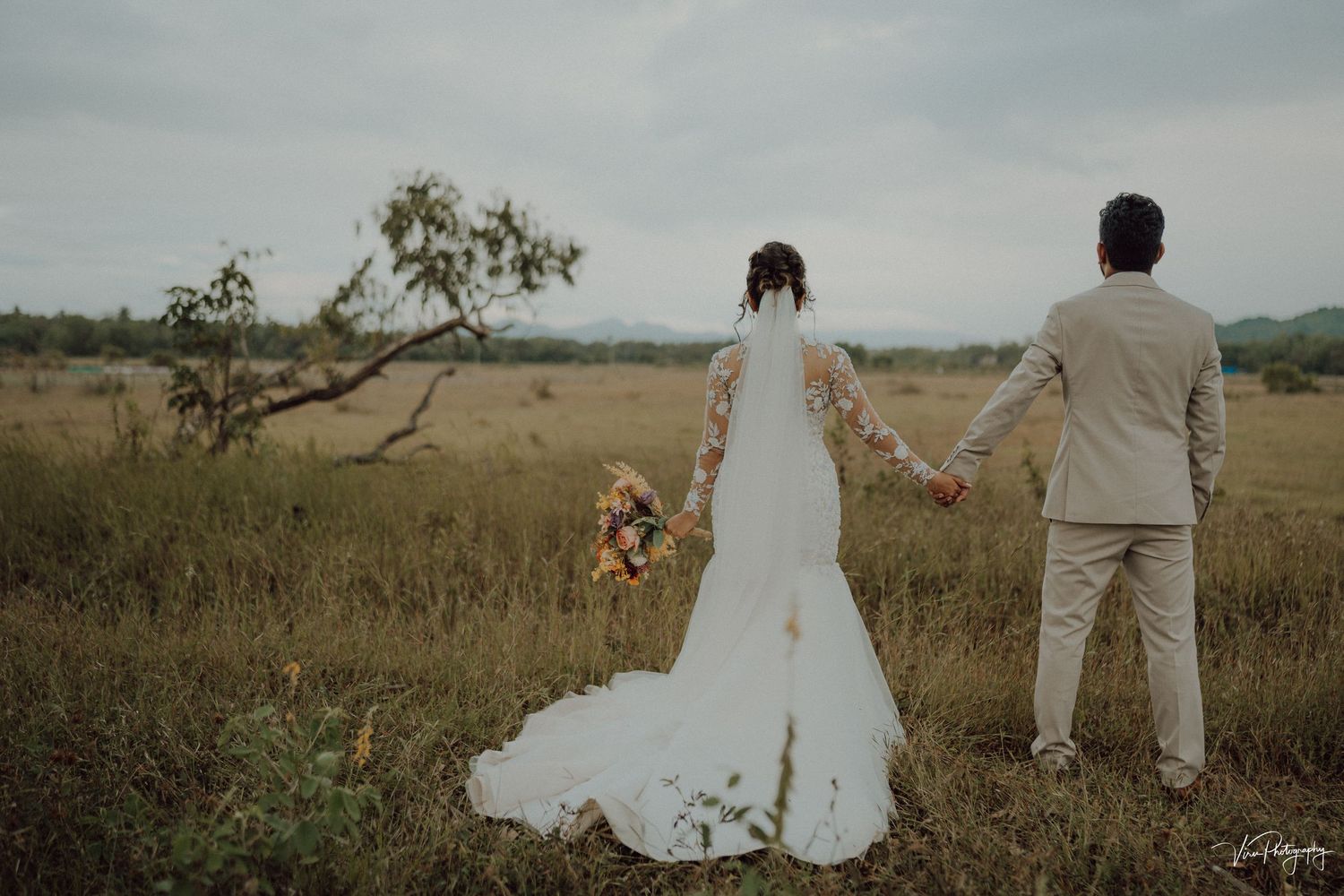 A romantic wedding couple walks hand in hand through a golden field at sunset with mountains in the distance.