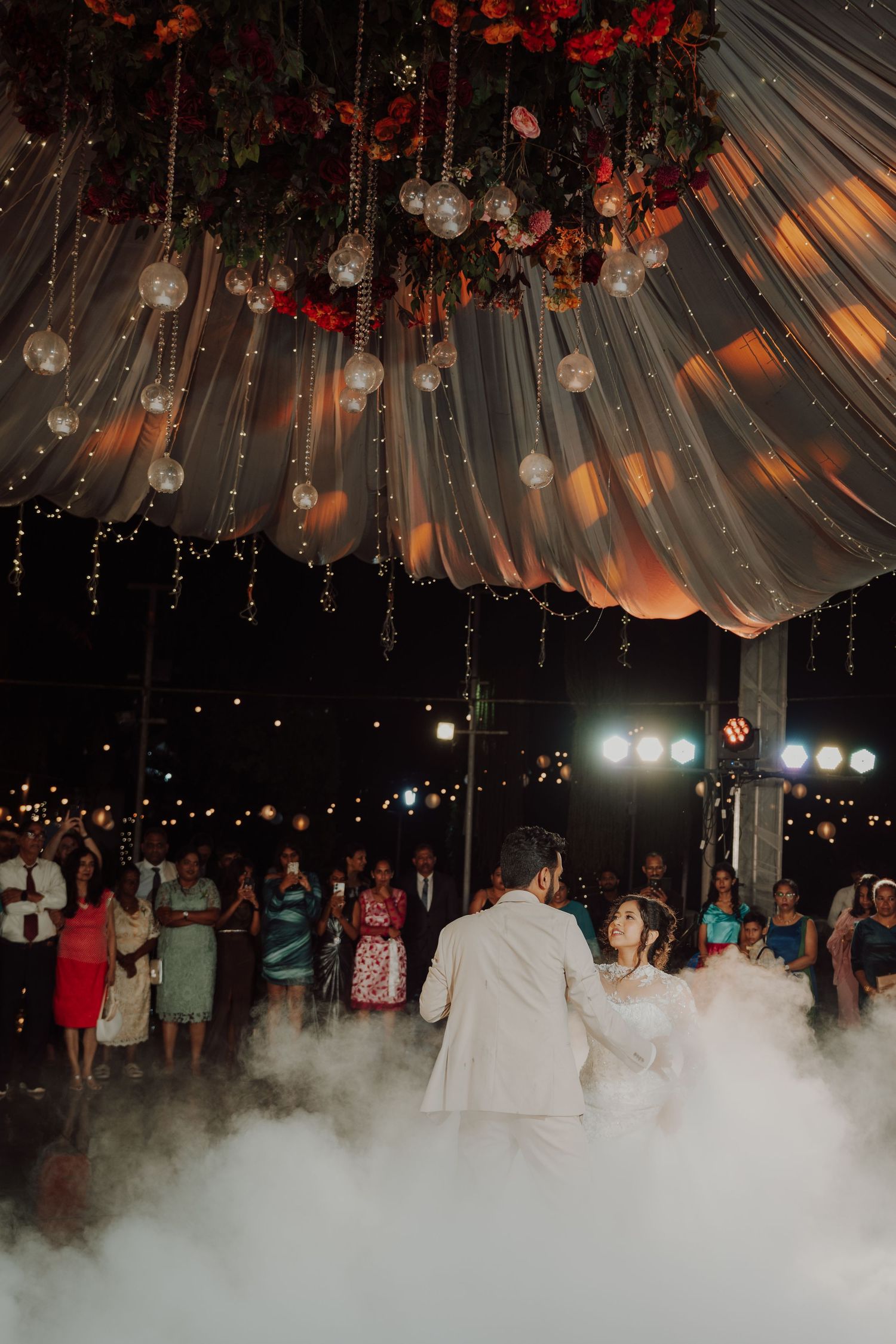 A bride and groom dance in a cloud of fog beneath a draped canopy with hanging flowers and glowing lights as guests watch.