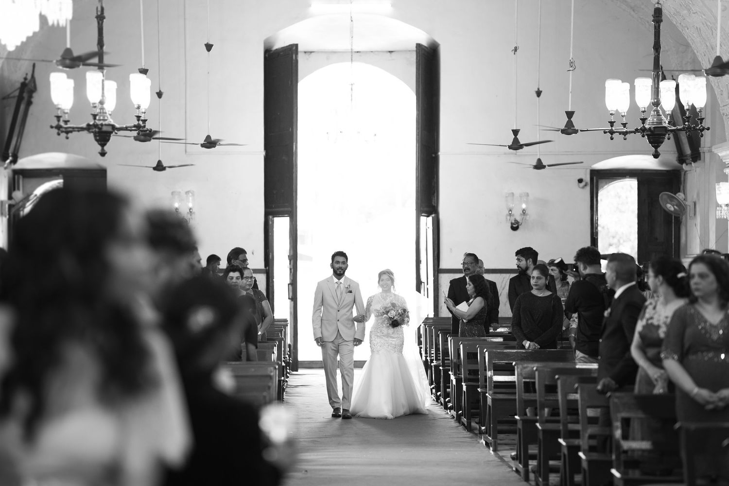 Bride and groom walk down the aisle of a church in black and white as seated wedding guests look on.