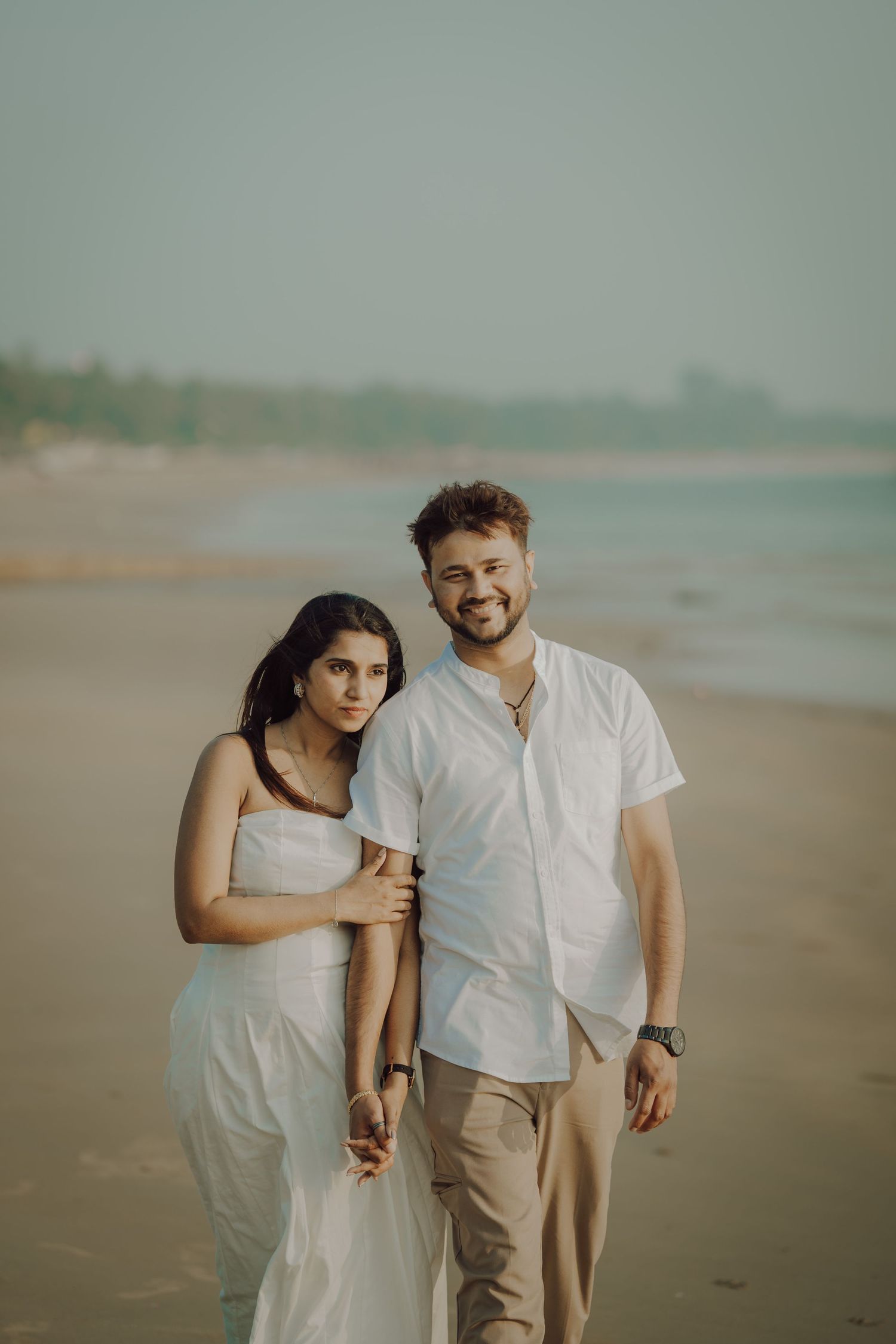 A couple in white outfits stands together on a misty beach during golden hour.
