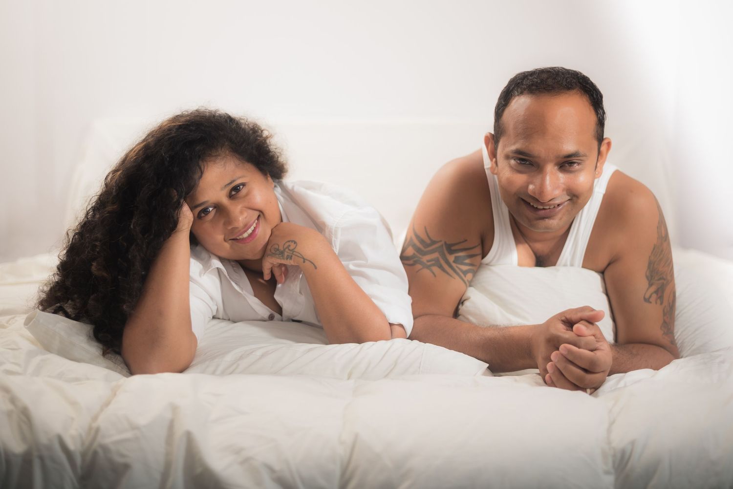 Two people lying on white bedding smile while relaxing together in casual attire.