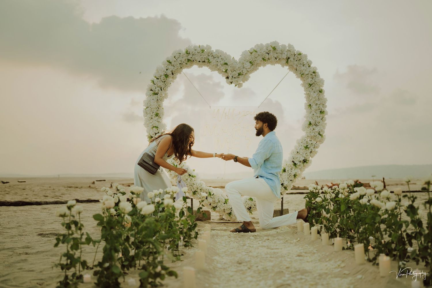 A romantic beach proposal scene with a floral heart arch against a misty coastal backdrop.