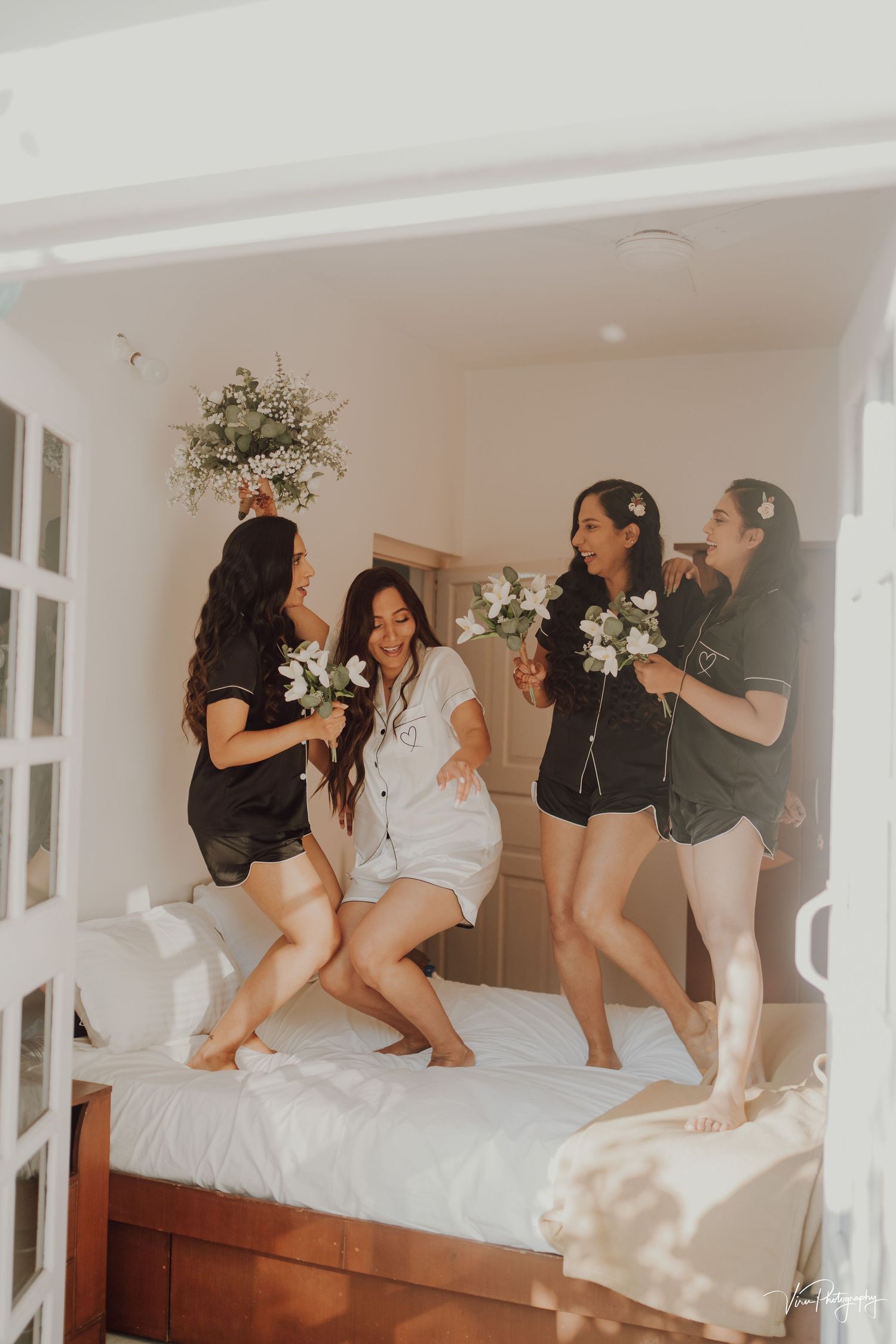 A group of friends in black and white outfits jump and dance together on a white bed while holding flowers.