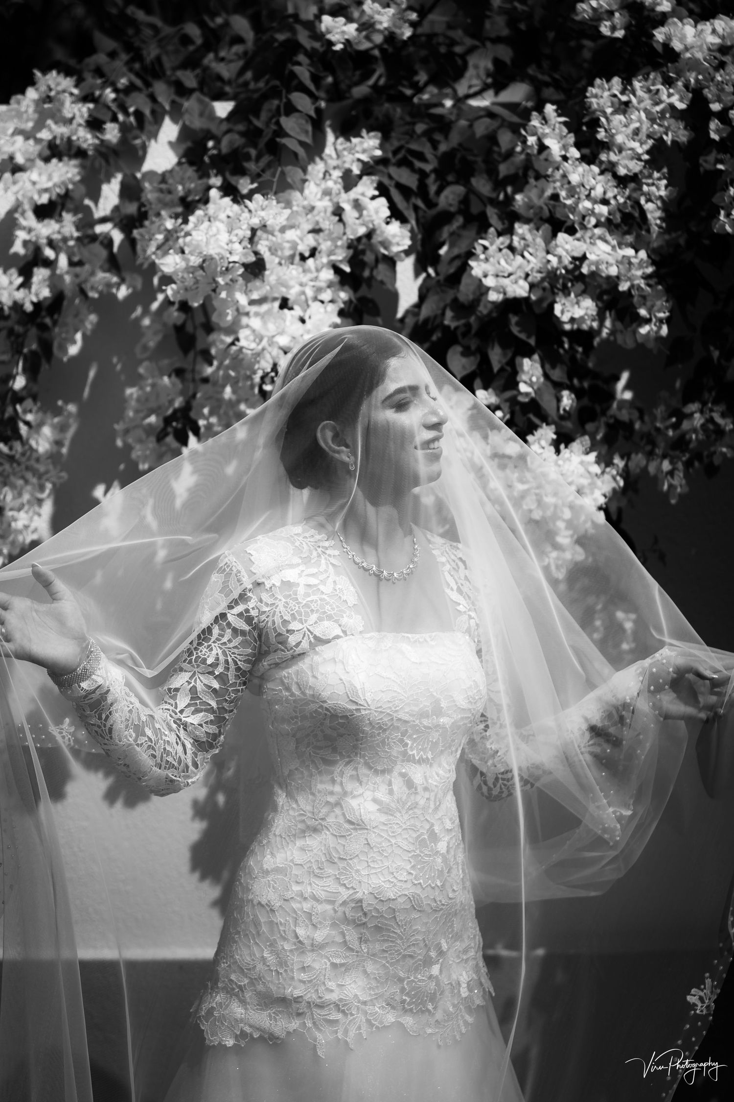 Ethereal black and white bridal portrait featuring delicate lace gown and flowing veil against flowering garden backdrop.