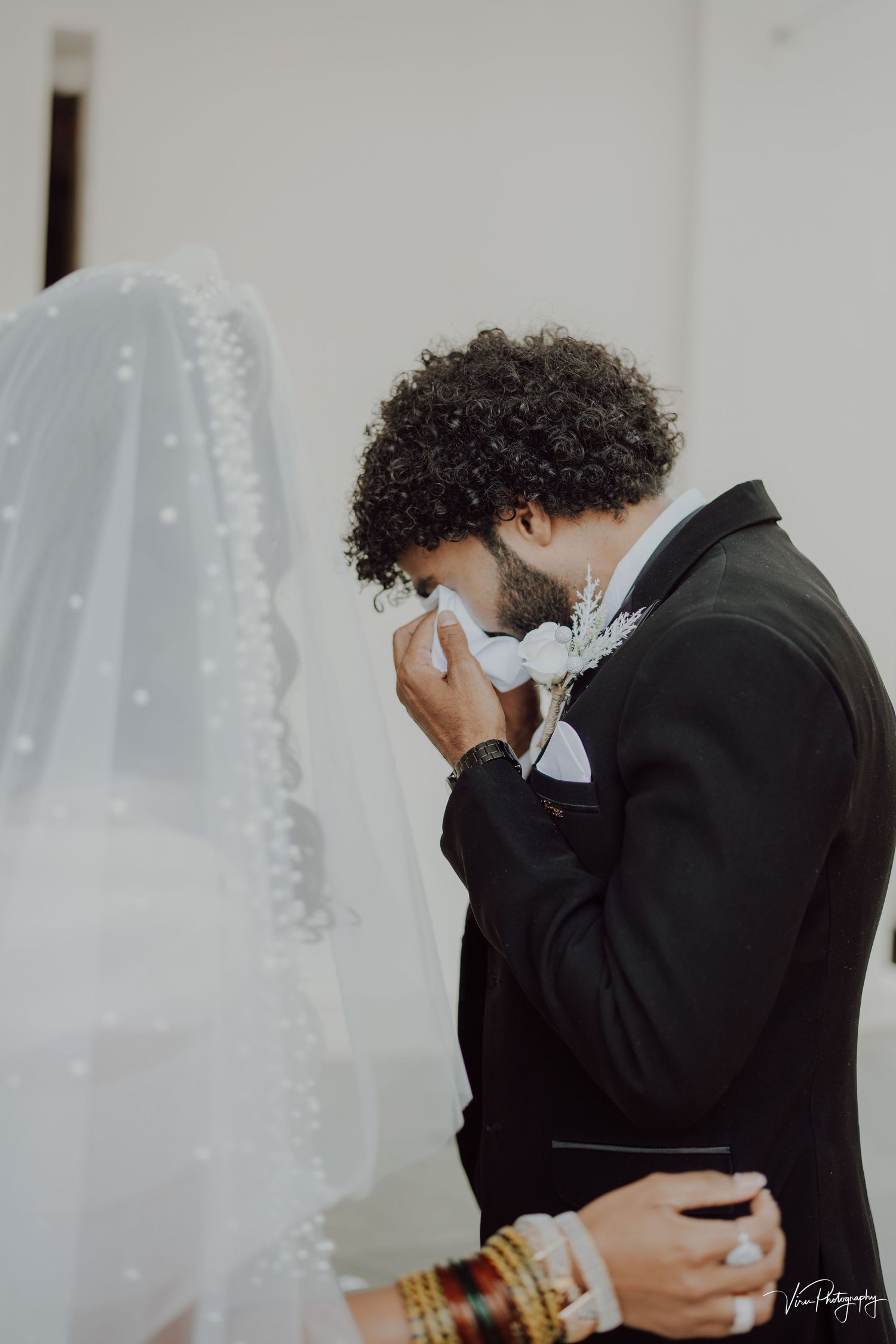 An emotional groom in a black suit wipes away tears as he sees his bride in her wedding dress with delicate veil details.