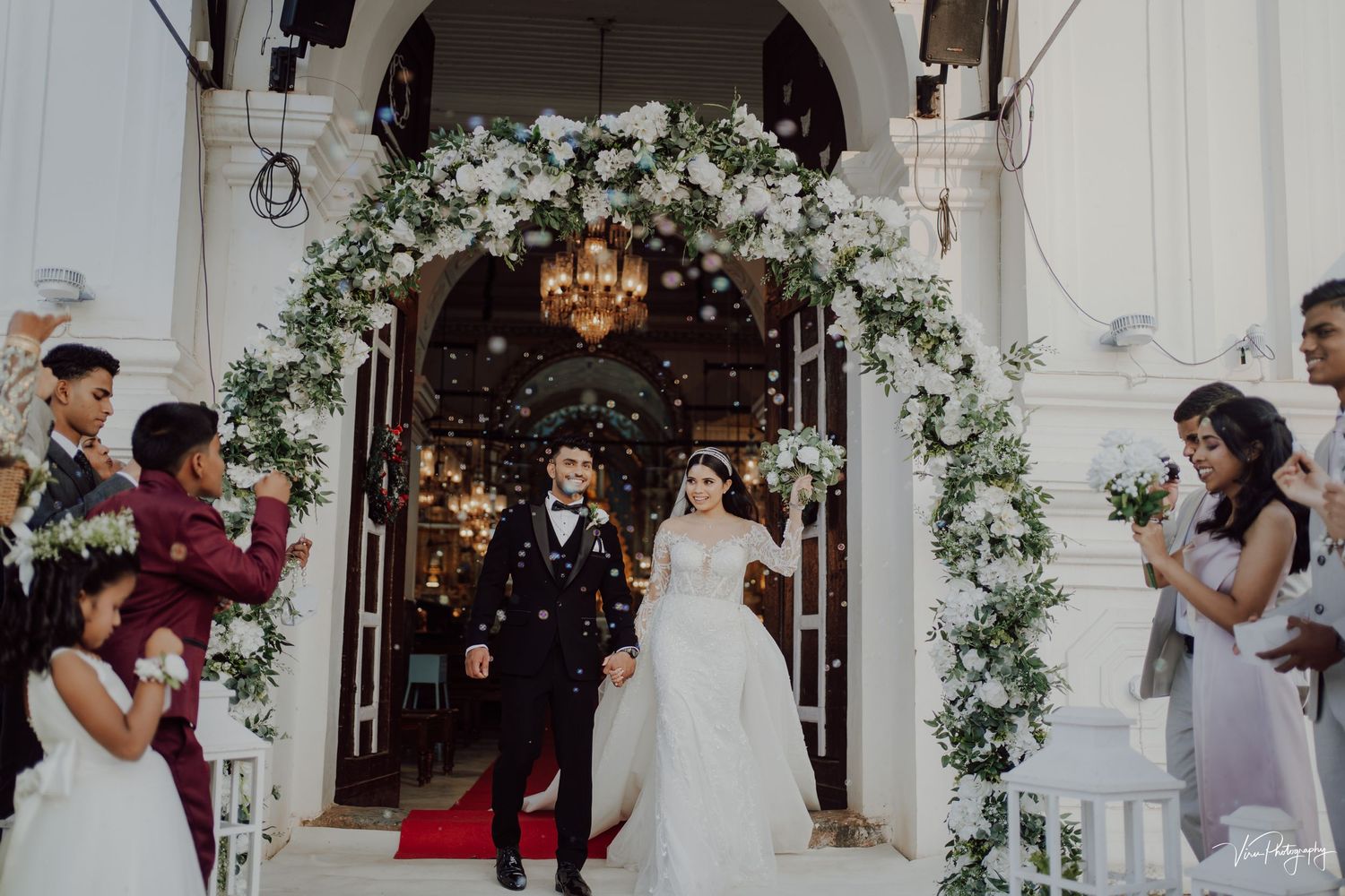 Wedding couple exits church under elegant white floral arch with chandelier and guests clapping.