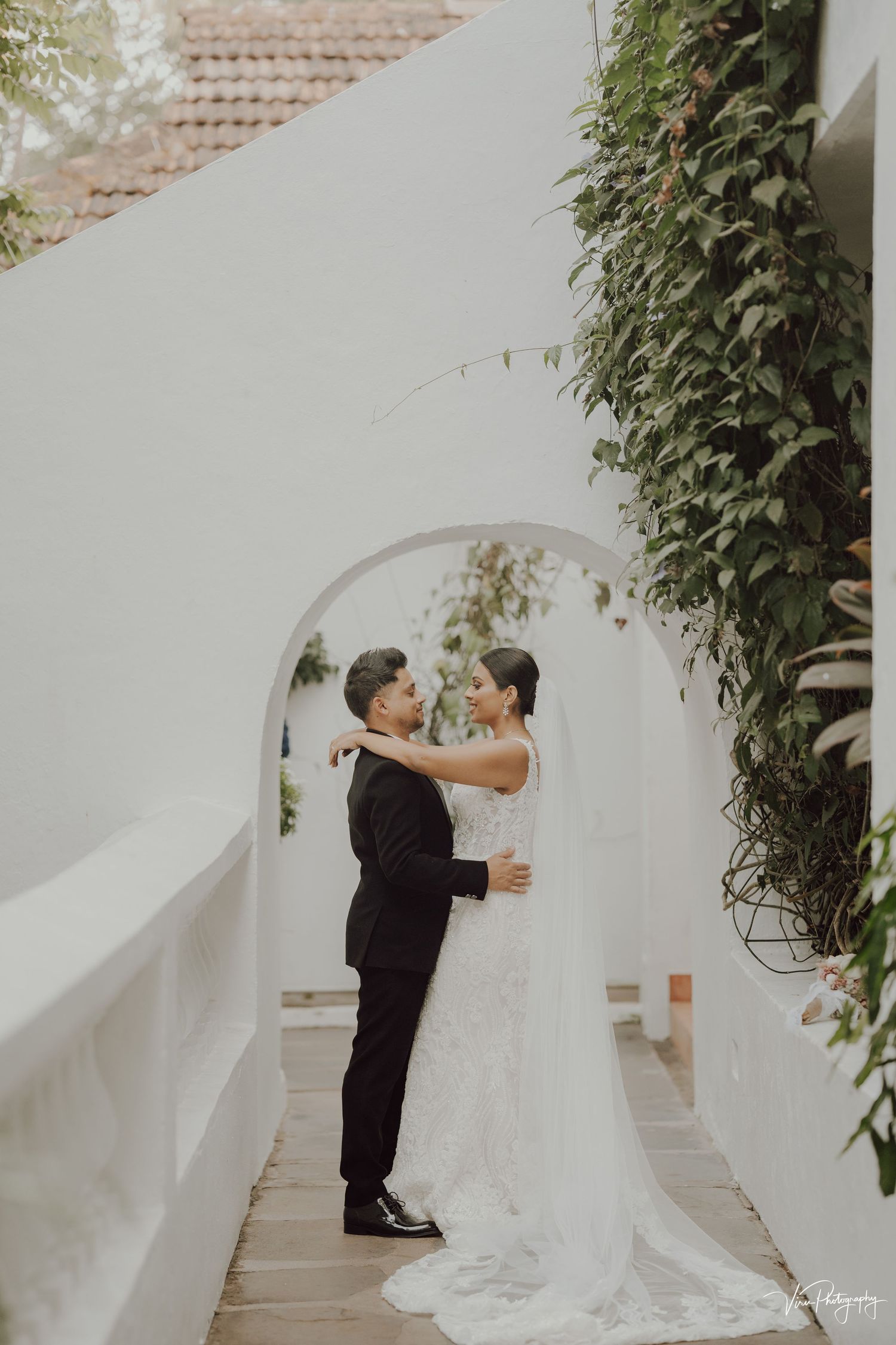 A couple embraces in a white archway with climbing vines framing the romantic Mediterranean-style architecture.