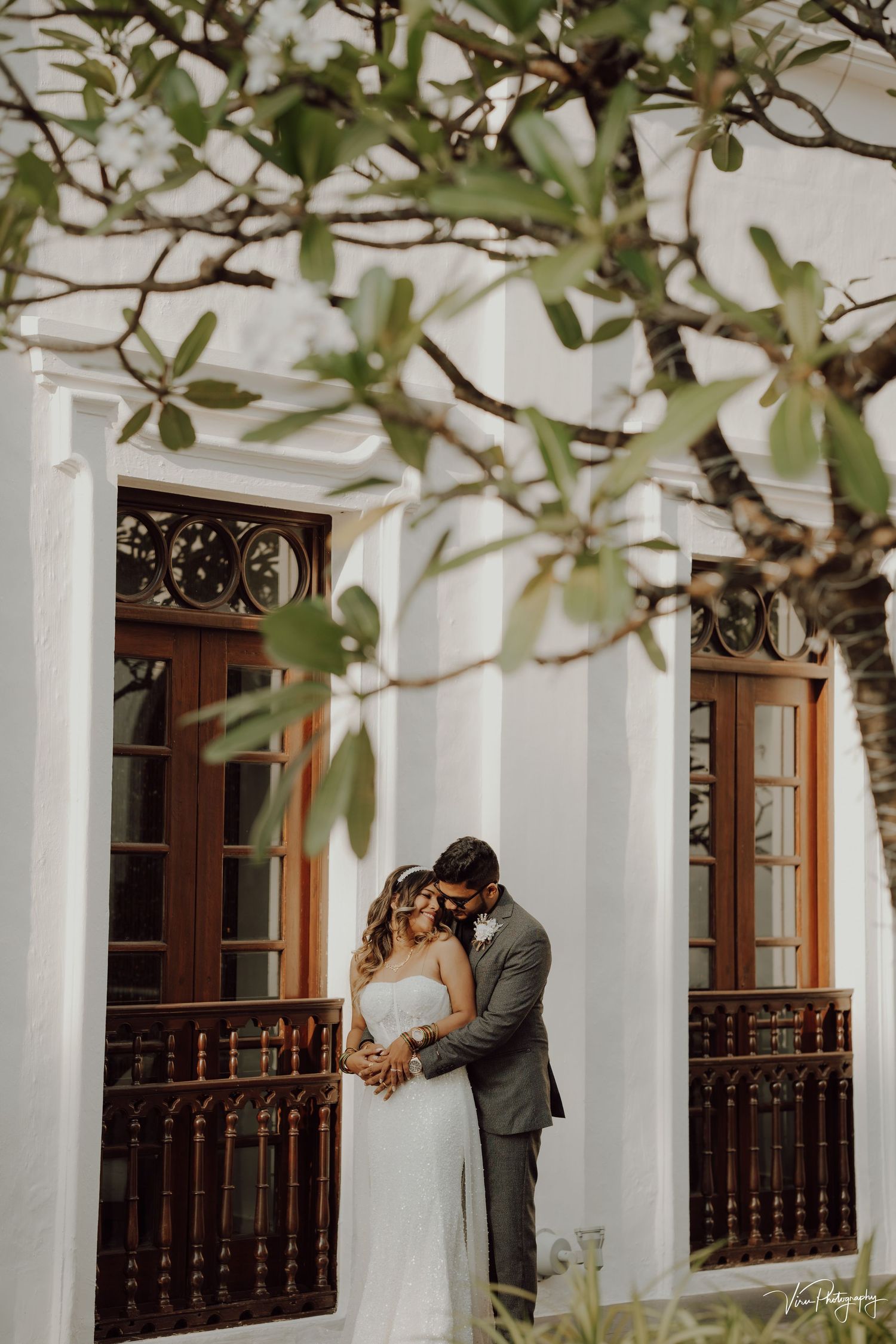 A couple shares an intimate moment on their wedding day framed by white columns and ornate wooden balustrades.