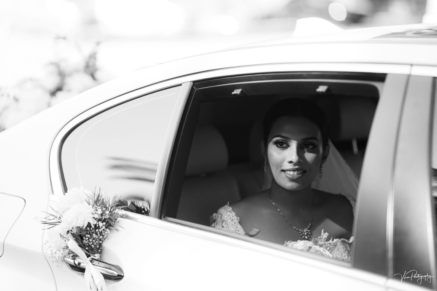 A black and white photograph shows an elegant bride sitting in the back of a car window on her wedding day.
