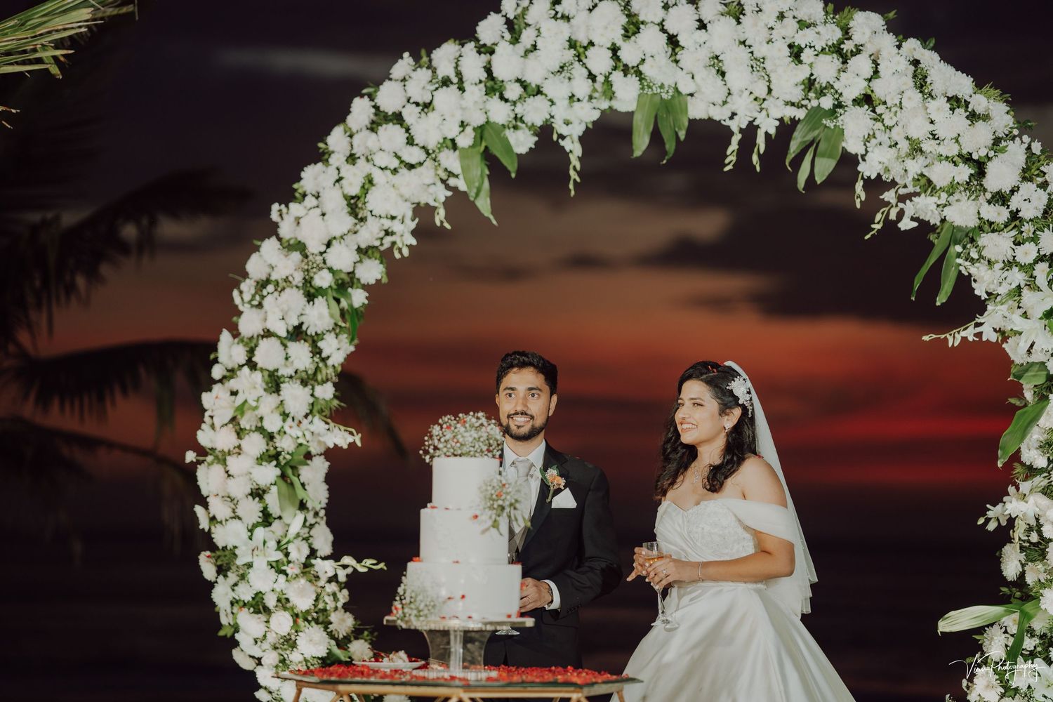 A wedding couple cuts their cake beneath an elegant white floral arch against a dramatic sunset sky.