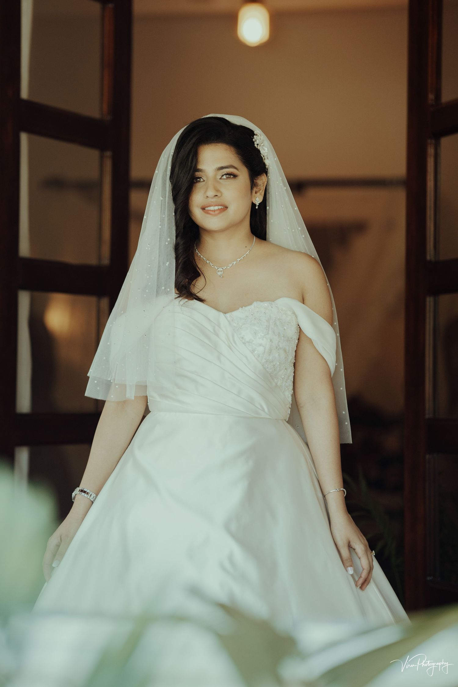 A bride in a classic white off-shoulder wedding gown and veil poses in warm lighting against a dark wooden doorway.
