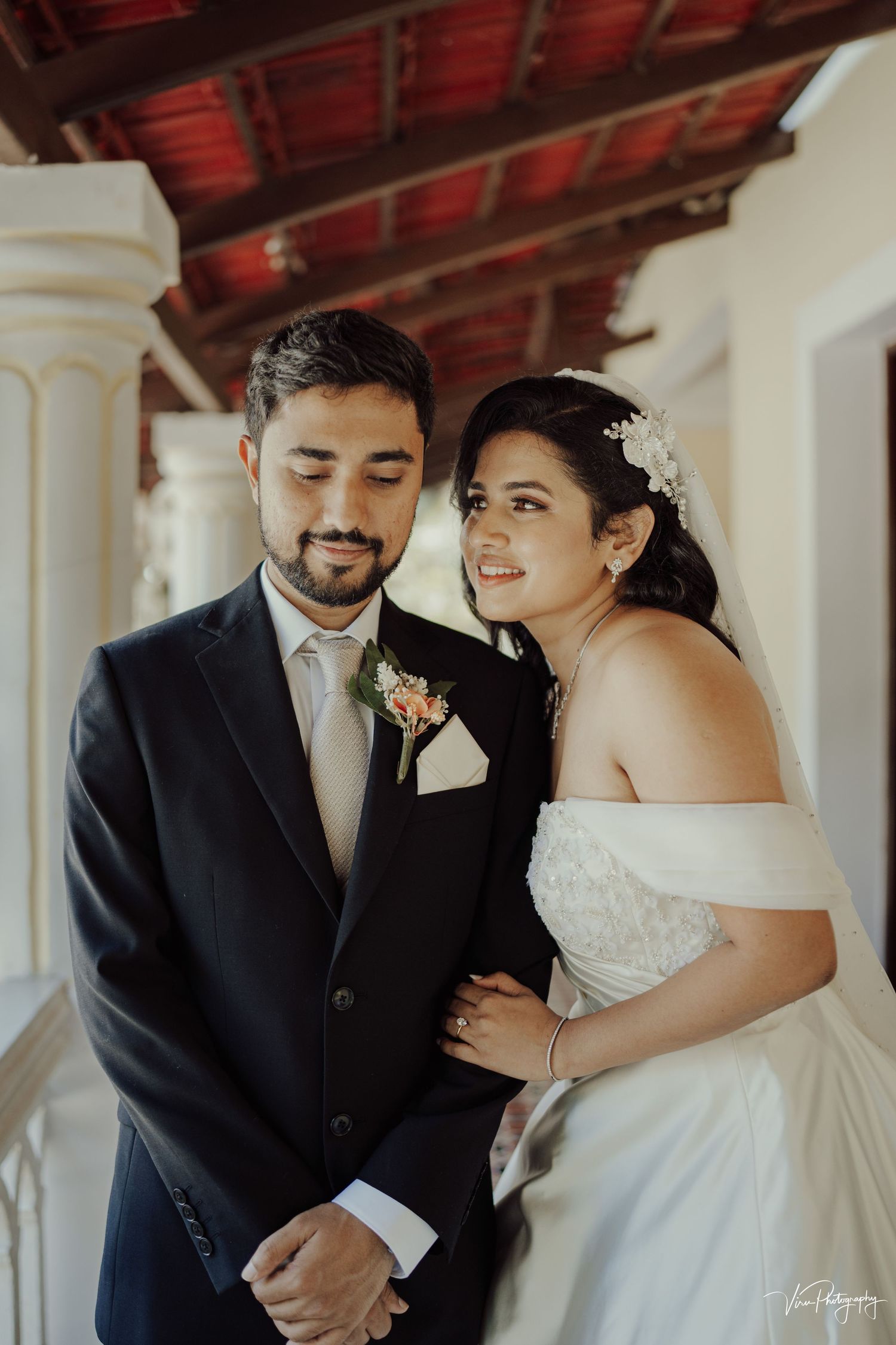 A couple in formal wedding attire poses together under a covered outdoor walkway with wooden beams overhead.