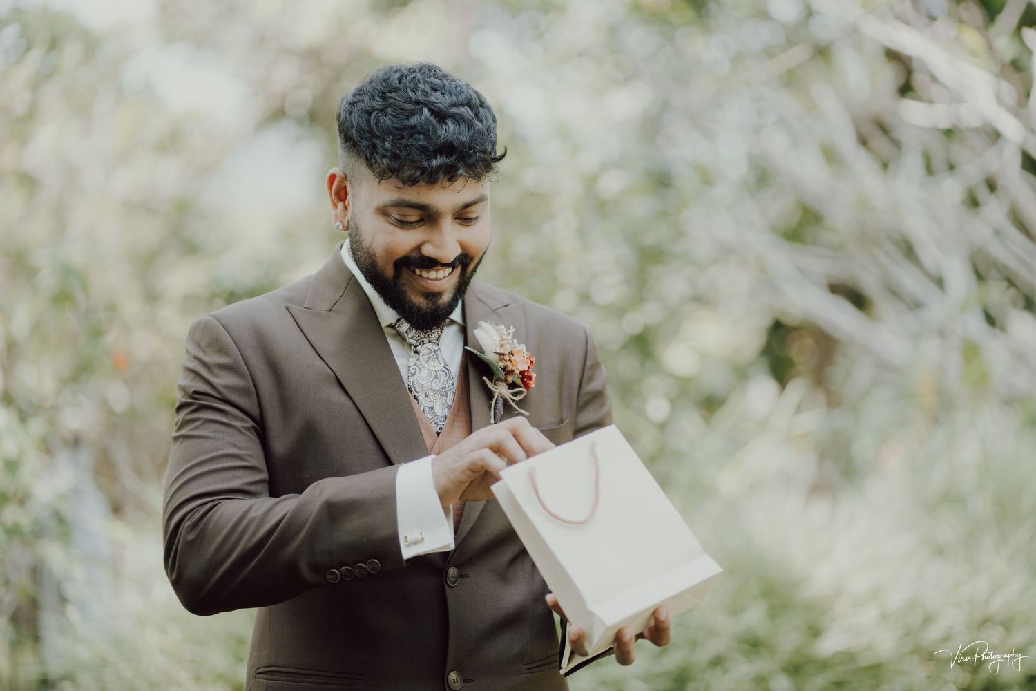 A groom dressed in a brown suit reads a personal letter while standing outdoors surrounded by white flowers.